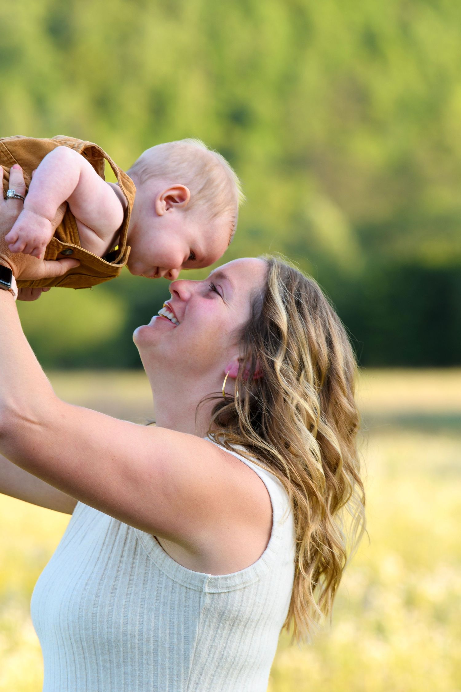 A joyful mother lifts her laughing baby high in the air in a sunny green field on a beautiful day.