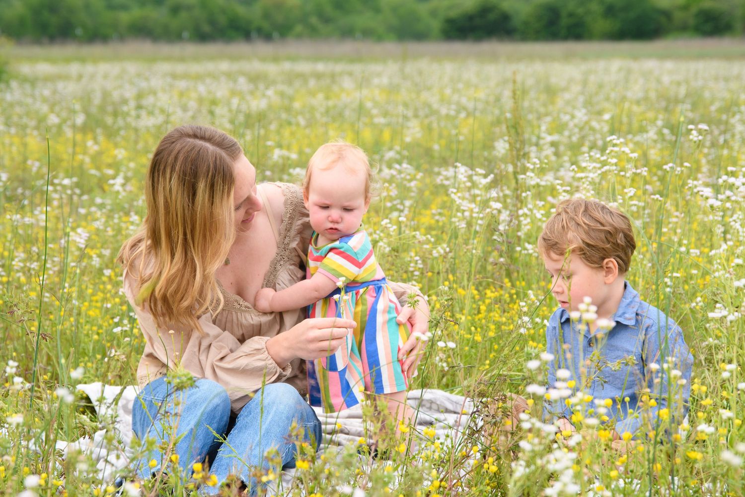 A mother and two children sit together in a bright wildflower meadow on a sunny day.