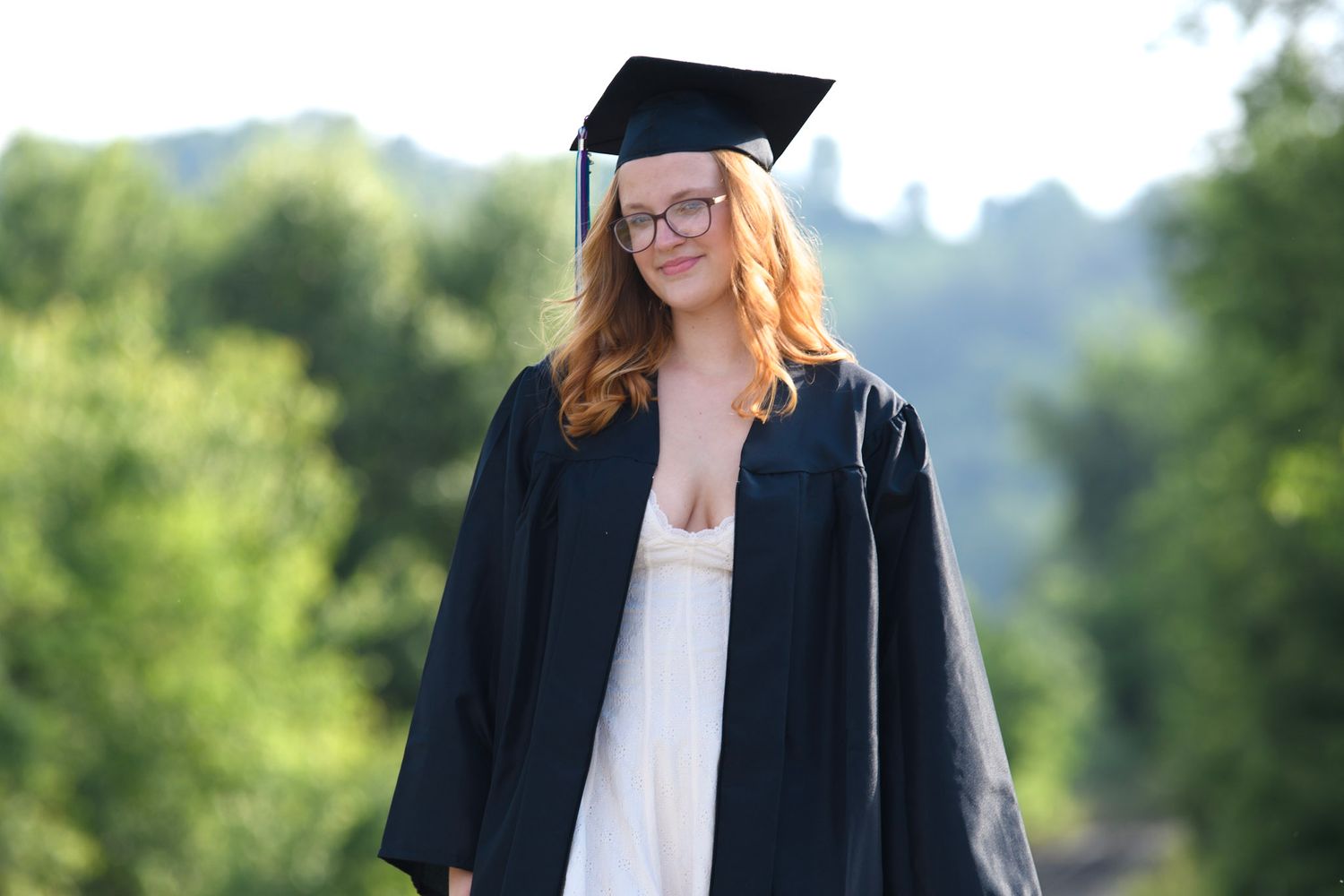 Young woman in black graduation cap and gown smiling outdoors with green trees in background.