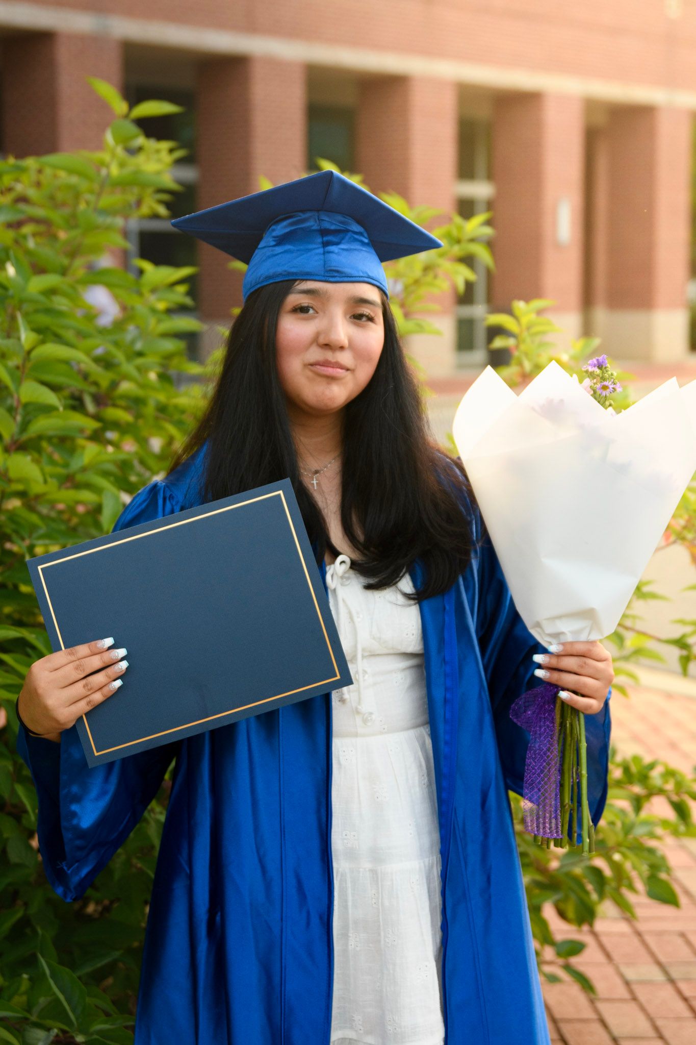 Young woman in blue graduation cap and gown holding diploma and flower bouquet outdoors.