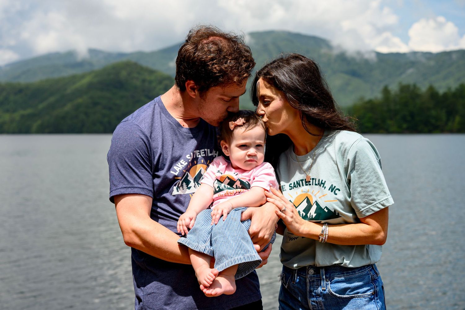 A couple kisses their baby by a mountain lake on a sunny day.