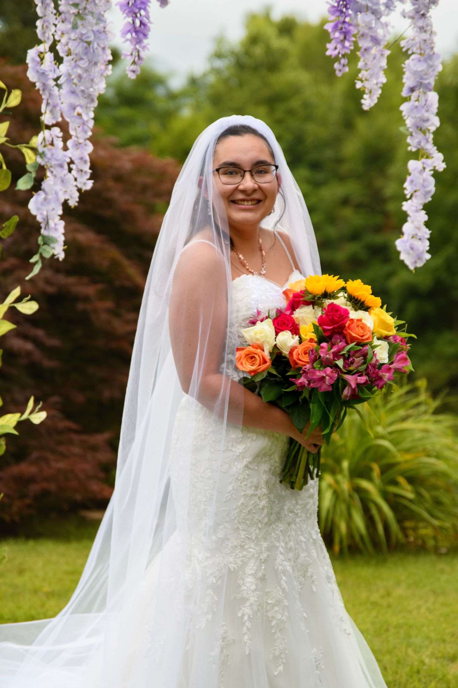 Smiling bride in white lace gown and veil holds colorful bouquet of roses outdoors under purple wisteria.