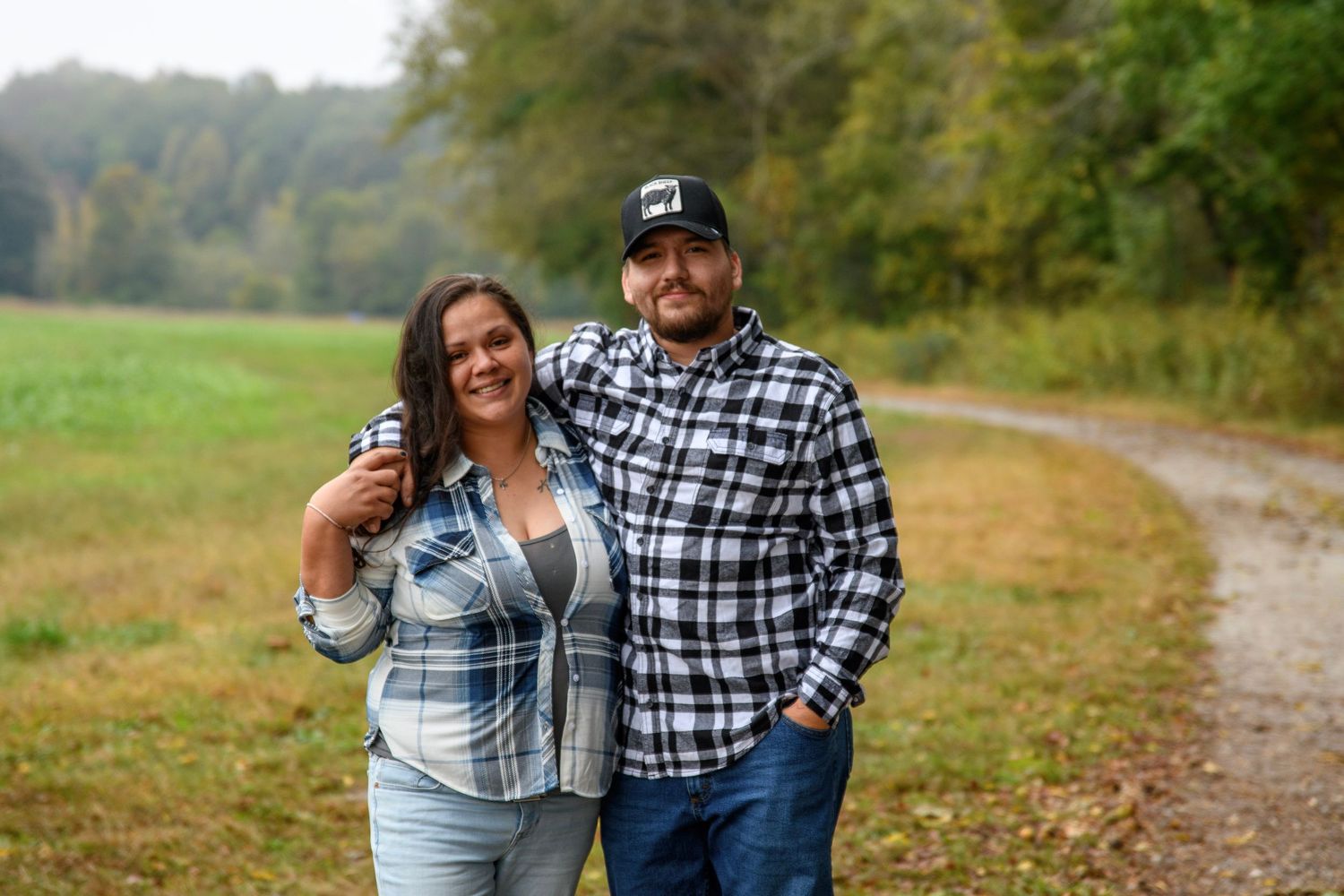 Smiling couple in matching plaid shirts pose together on a tree-lined country path.