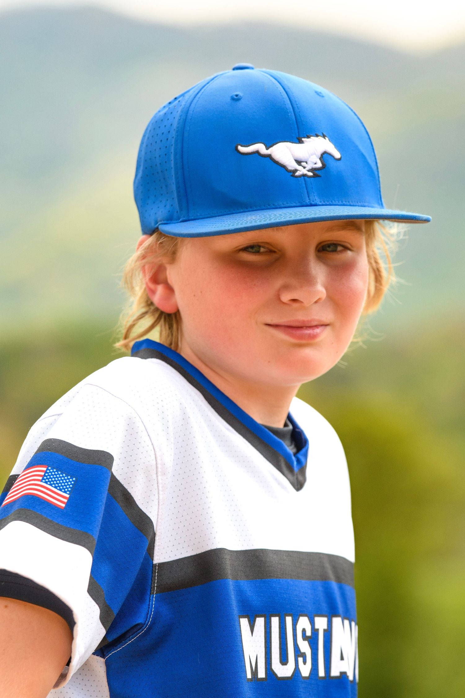 Close-up portrait of young blonde baseball player in blue Mustangs cap and jersey with mountain background.