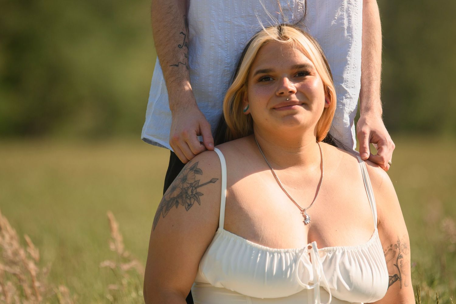 Blonde woman with tattoos in a white top smiling softly in a sunlit field with tall grass.