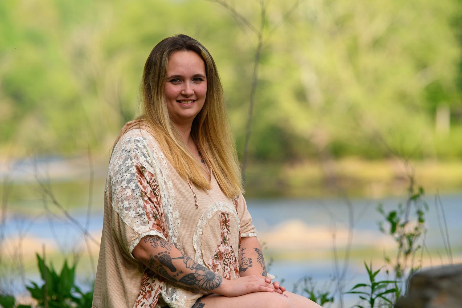 Smiling tattooed woman in lace top sitting outdoors near river surrounded by spring greenery.