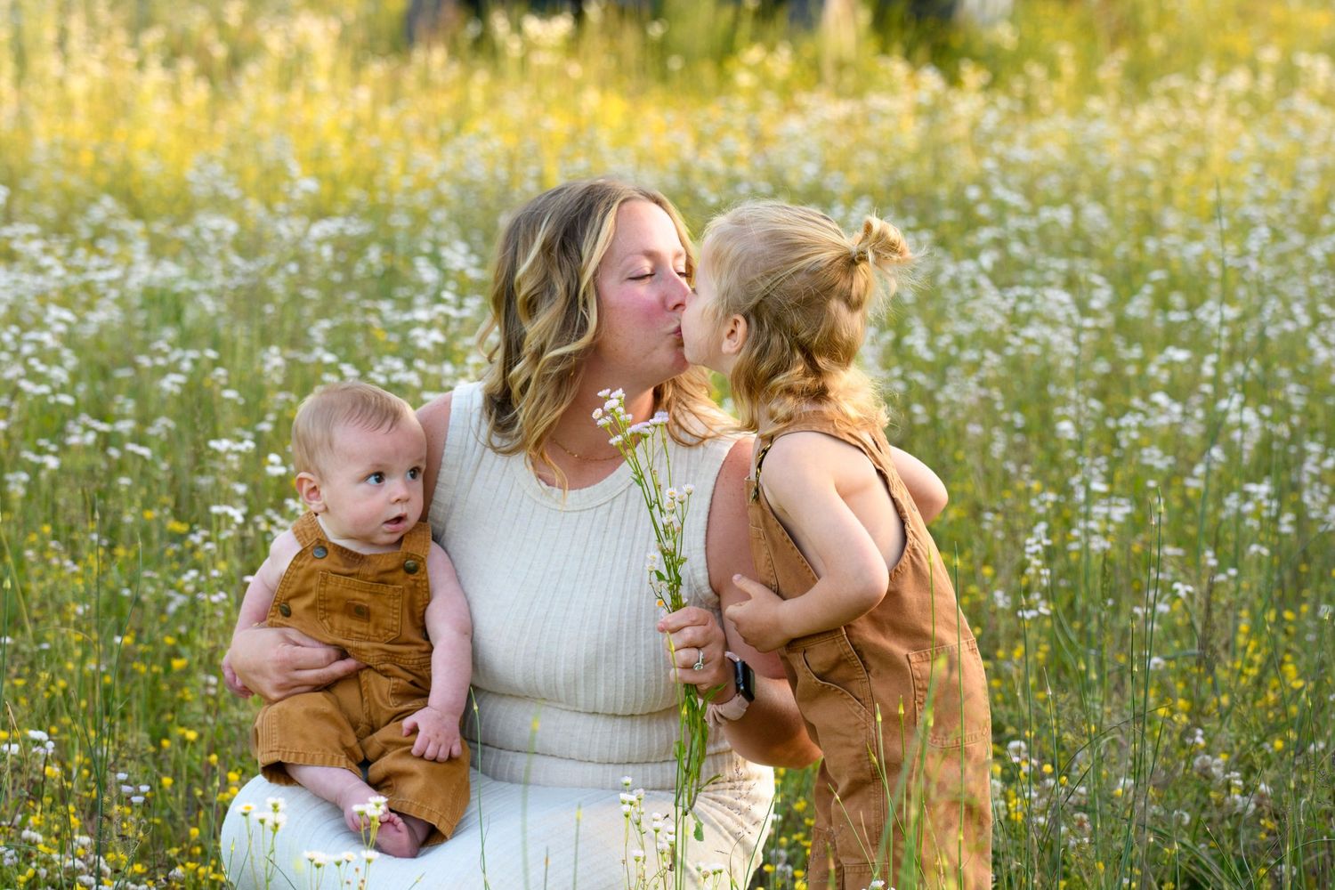 Mother in white dress holding baby while toddler kisses her cheek in a golden wildflower field.