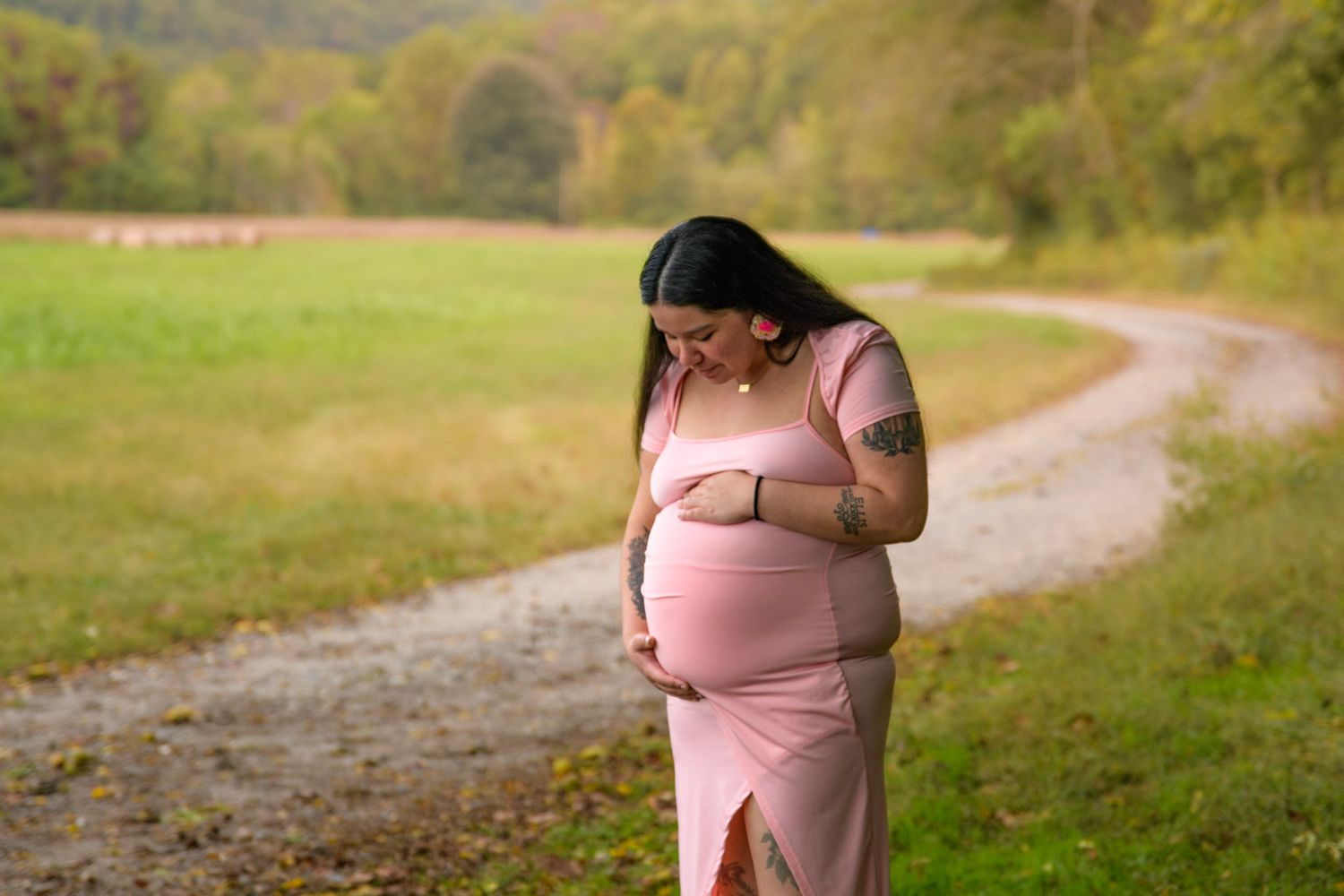 Pregnant woman in pink dress cradling baby bump outdoors on scenic country path surrounded by lush green fields.