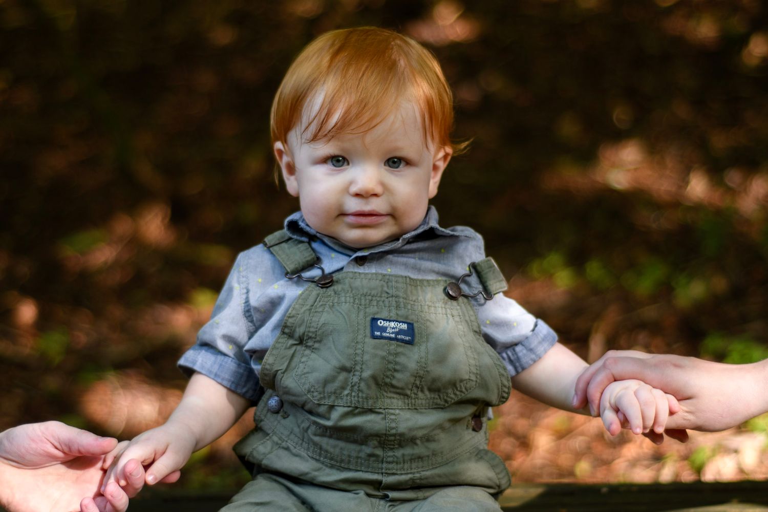 Red-haired toddler in olive OshKosh overalls sitting outdoors, held by adults hands on a wooden bench.