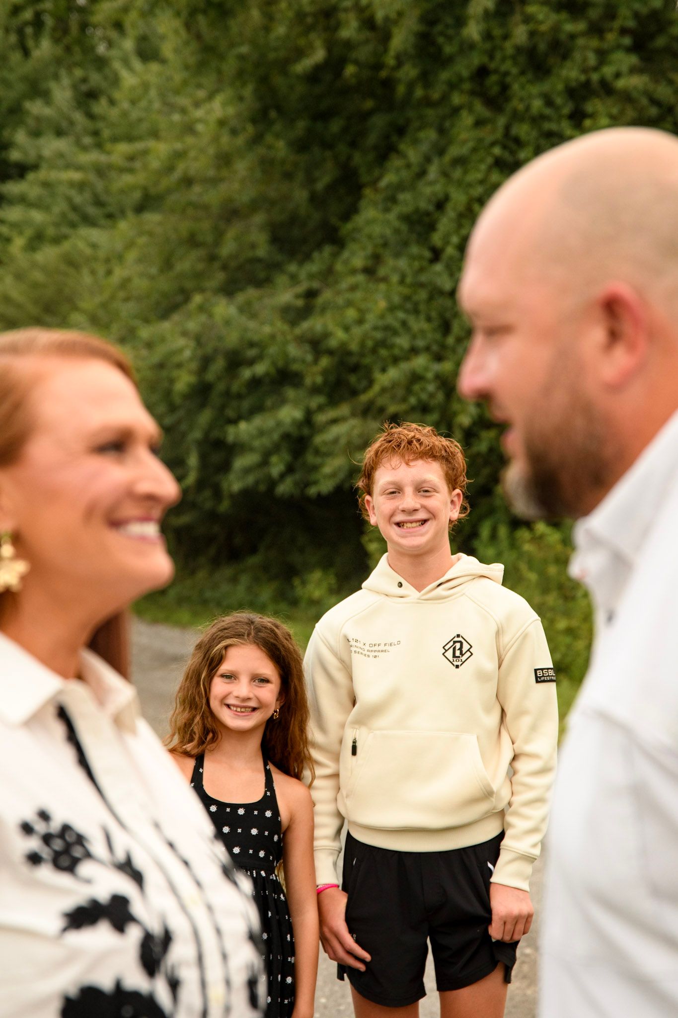 A happy family of four smiling outdoors, with two adults in foreground and two children in background near green trees.