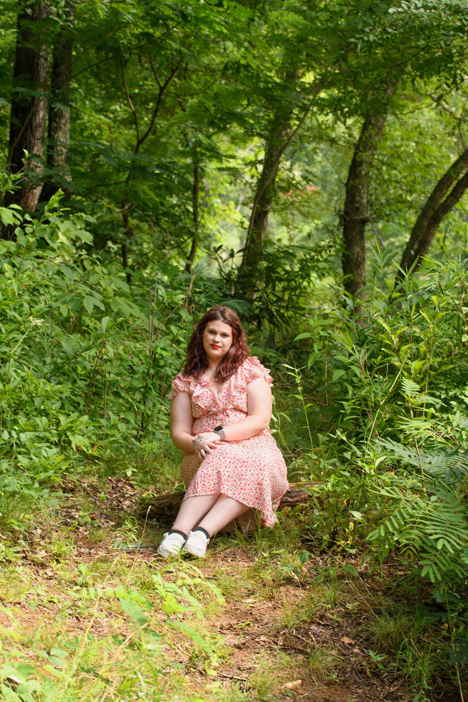 Young woman in floral pink dress sitting on log in lush green forest setting.