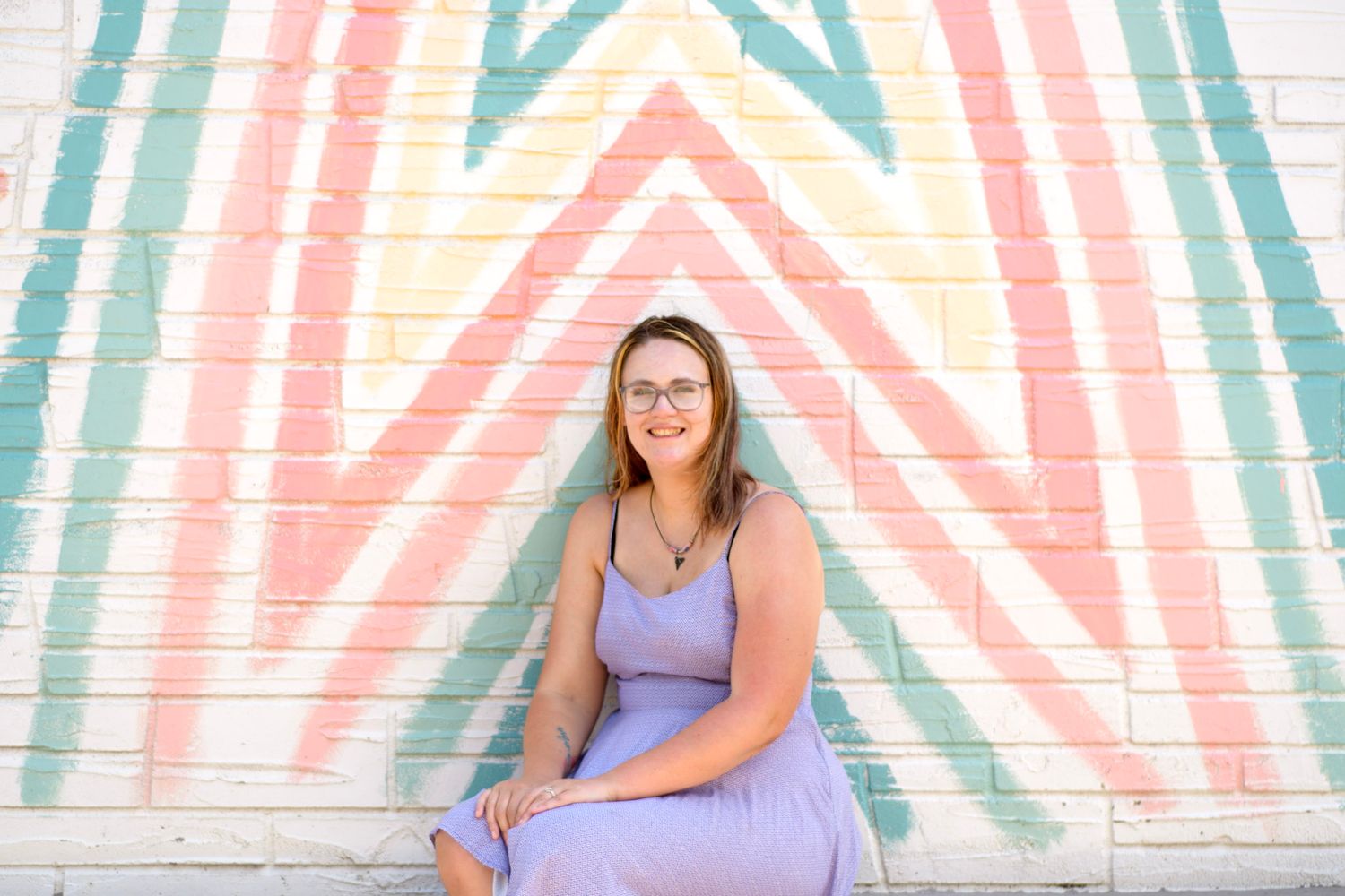 Smiling woman in lavender dress sitting against colorful rainbow chevron mural wall.