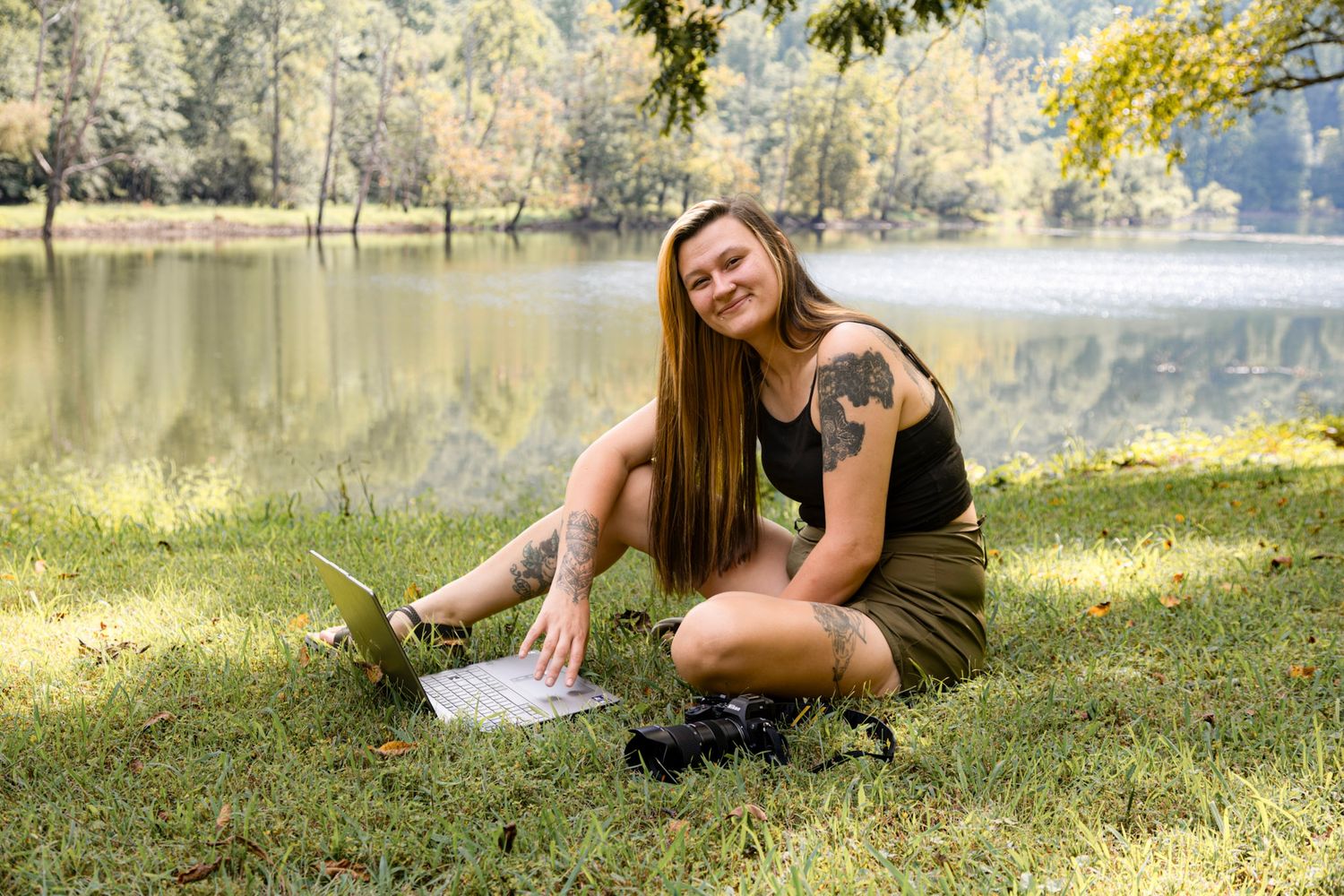 Young woman with tattoo sitting on grass by a lake, smiling at camera with laptop, enjoying outdoor remote work.