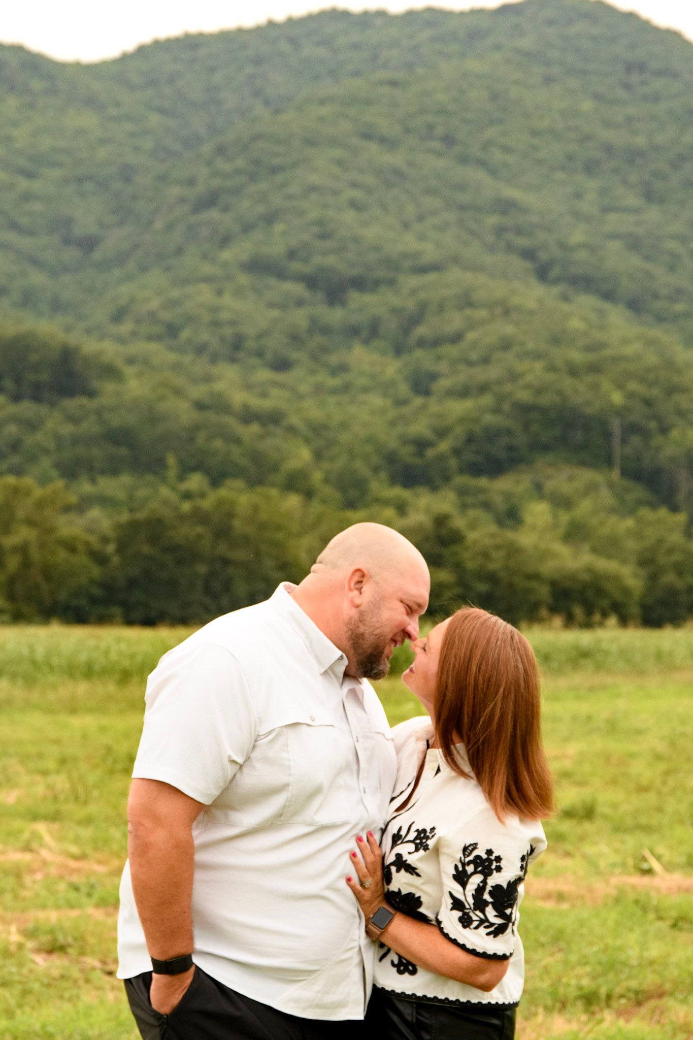 Couple sharing a romantic moment in a green field with lush mountain backdrop, man in white shirt embracing woman in black and white top.