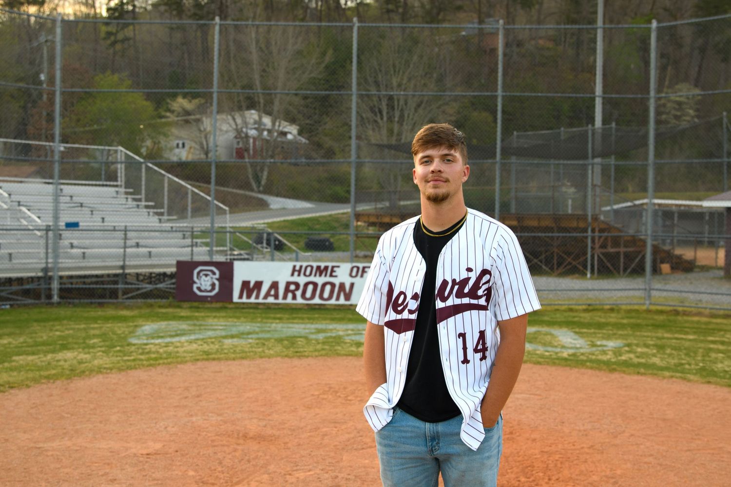Teen boy in baseball jersey number 14 stands on a baseball field in front of a Maroons sign.
