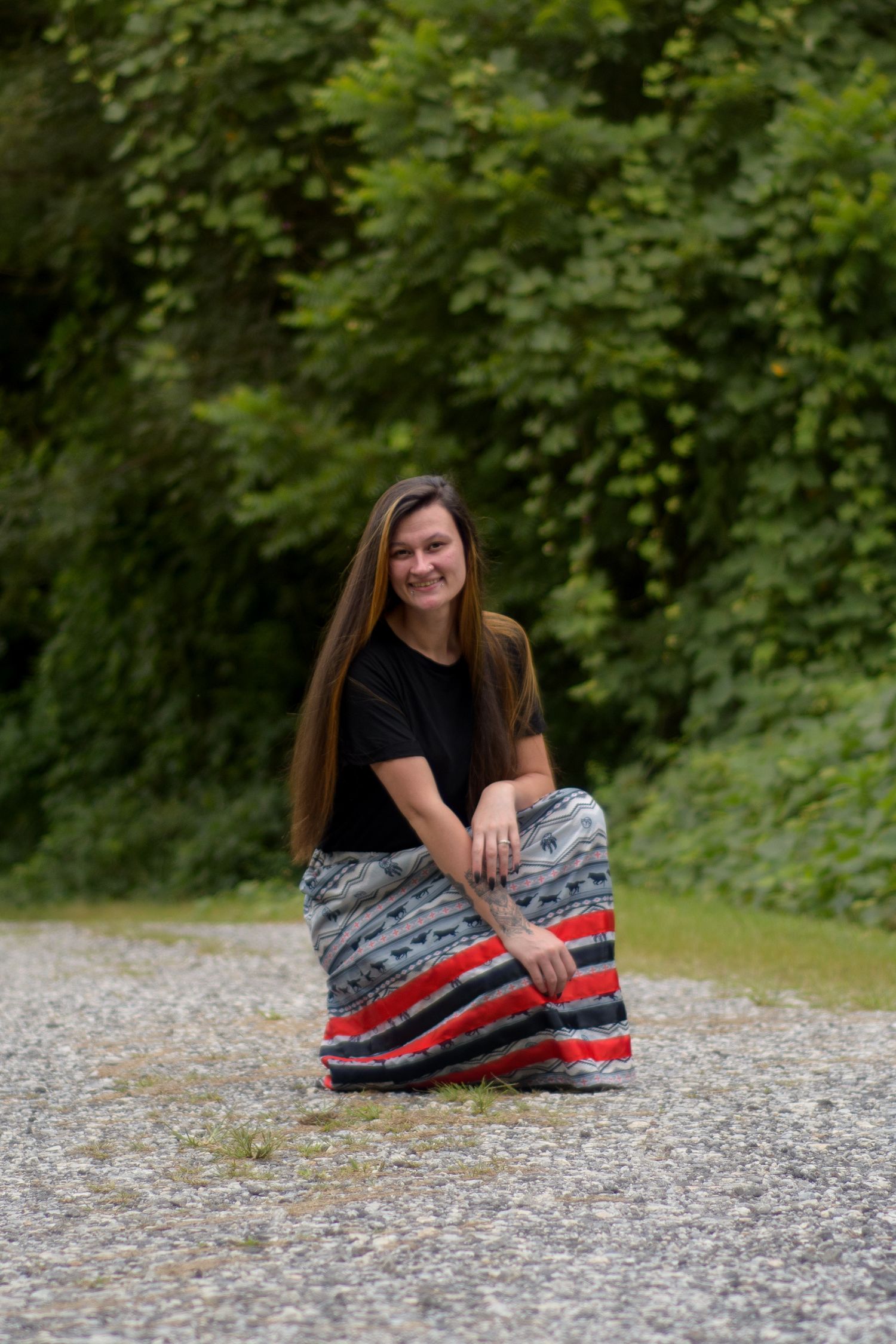 Smiling young woman with long brown hair crouching on a gravel path surrounded by lush green trees.