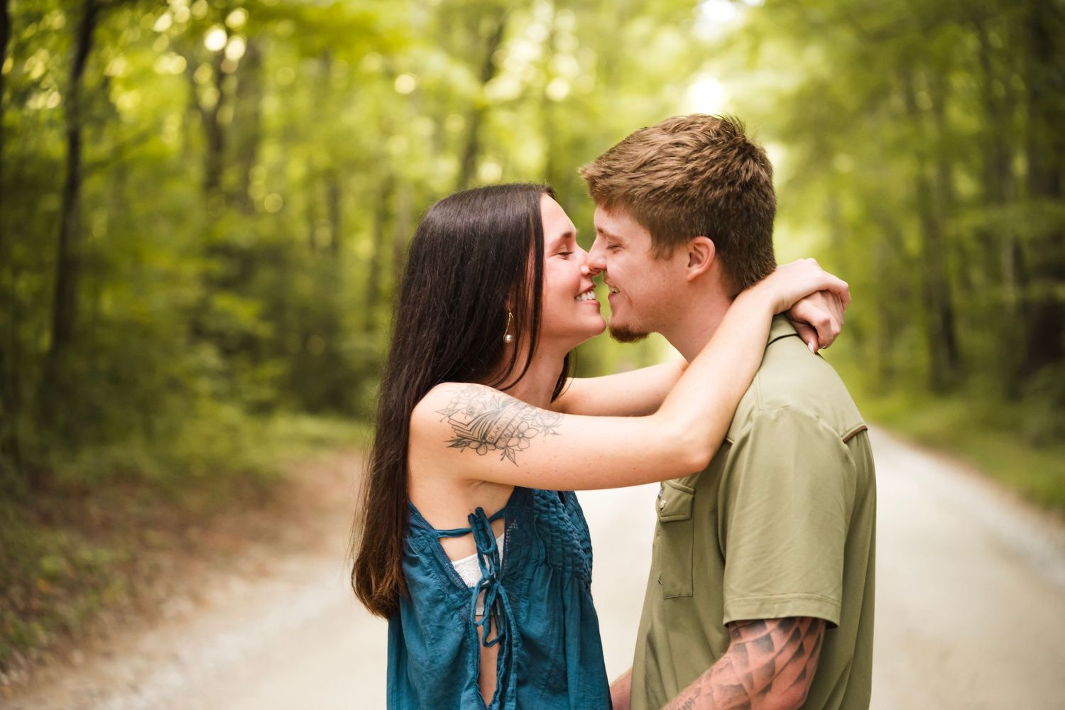 A couple shares a romantic moment on a forest path, the woman with a tattoo embracing the man in a green shirt.