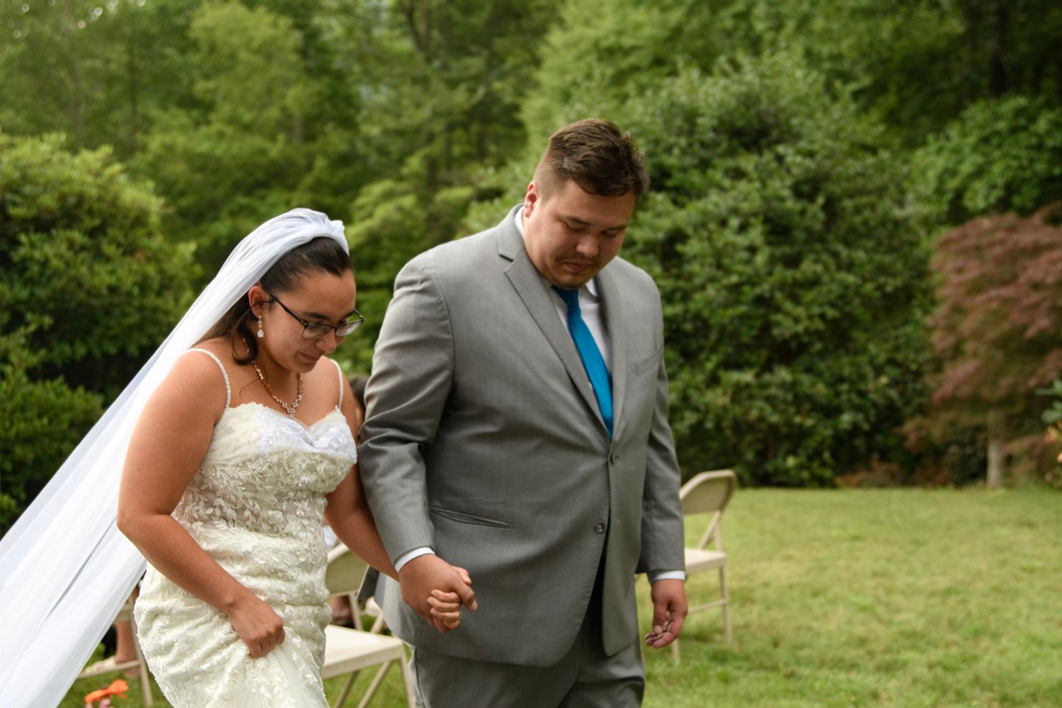 Bride in white gown and veil walks with groom in gray suit and teal tie during outdoor wedding ceremony.