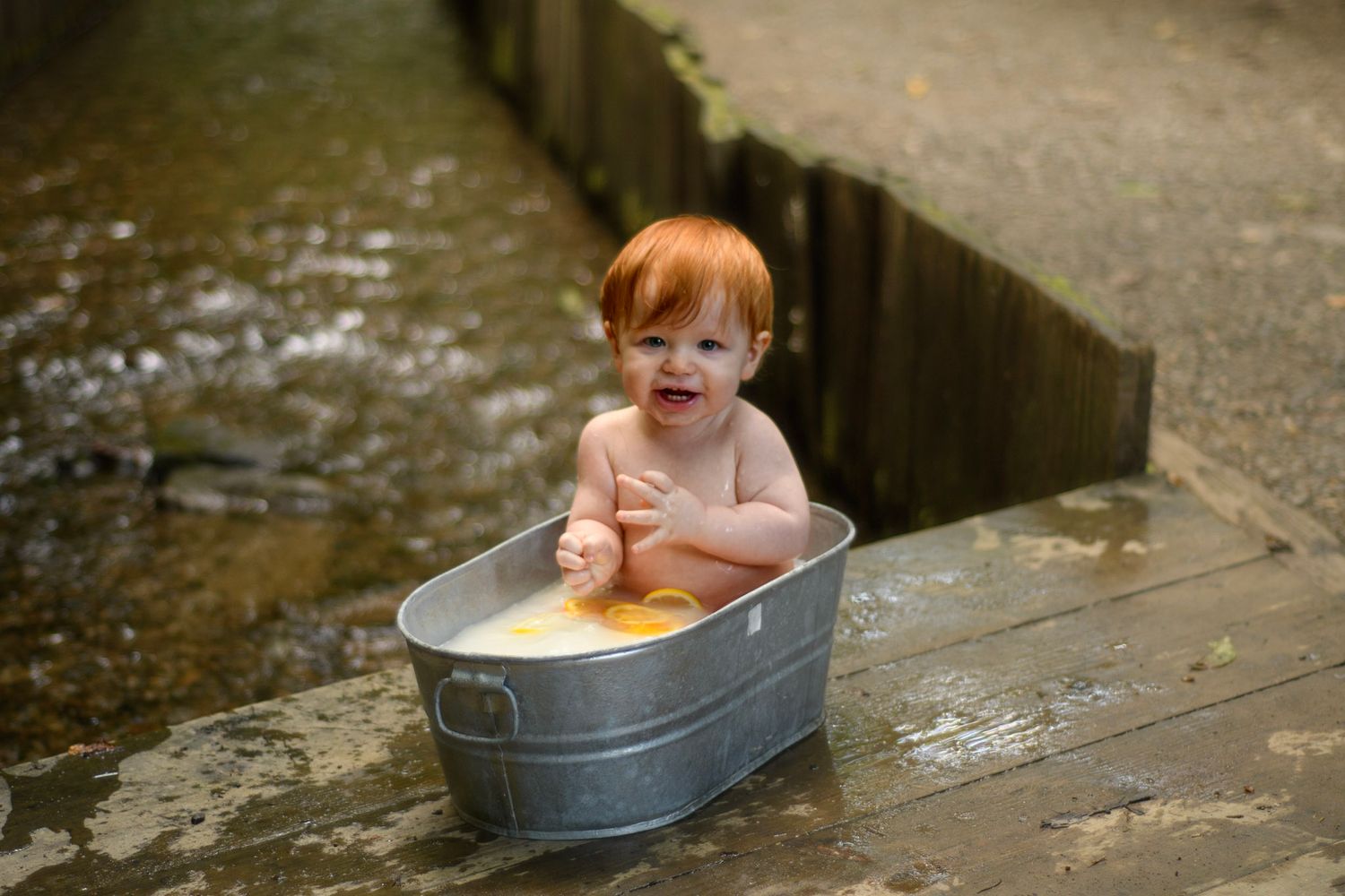Red-haired baby sitting in a metal tub outdoors near water, smiling and playing, in a charming rustic setting.