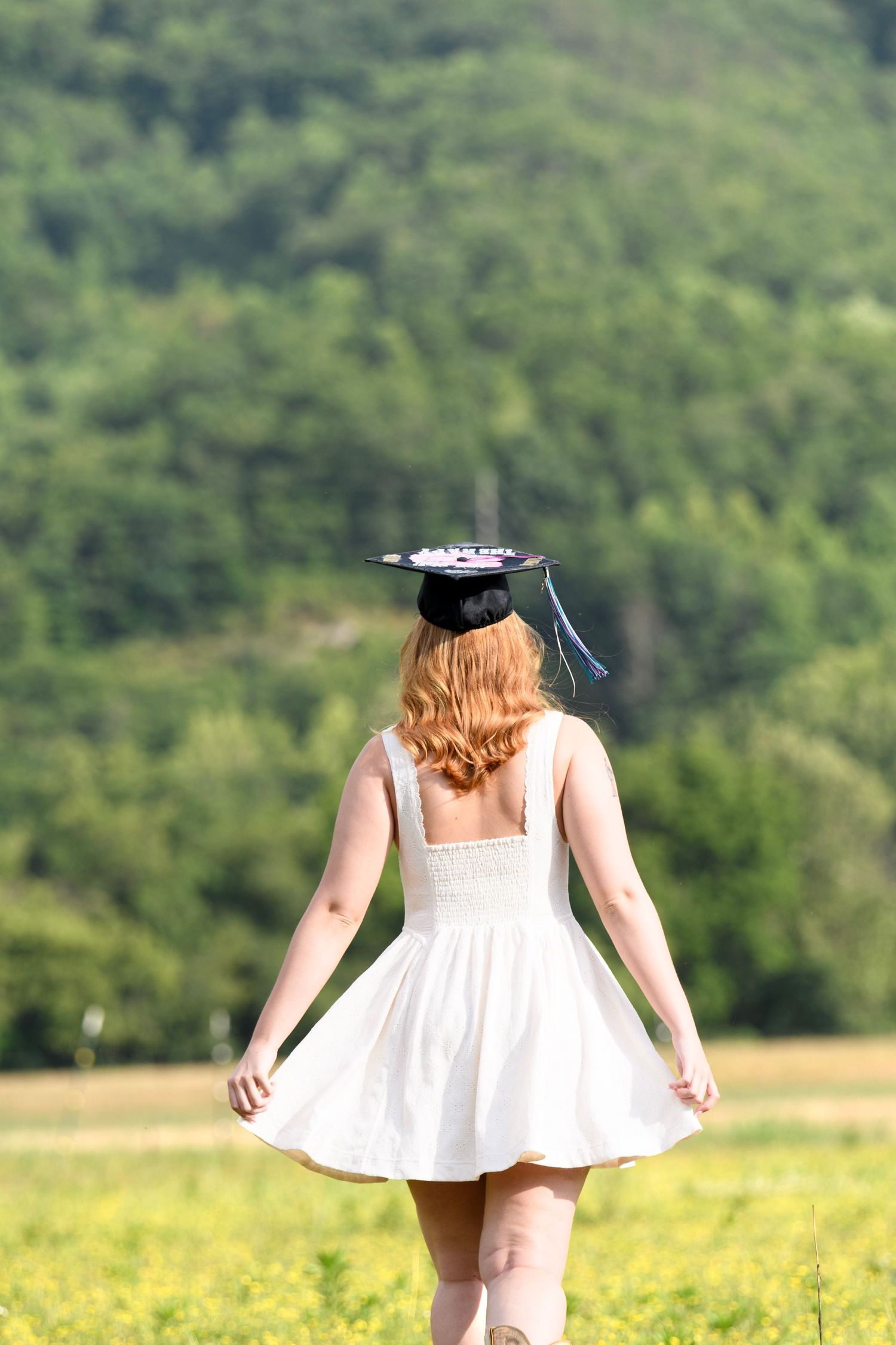 Red-haired graduate in white dress wearing mortarboard cap walks through sunny green meadow, viewed from behind.