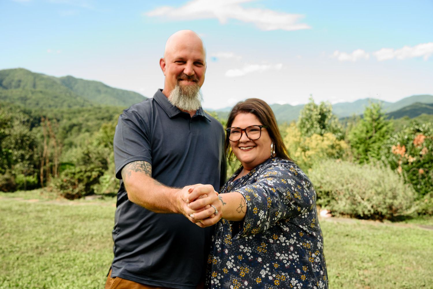 Smiling bearded man and woman with glasses clasp hands outdoors on a grassy lawn with green mountains and blue sky behind them.