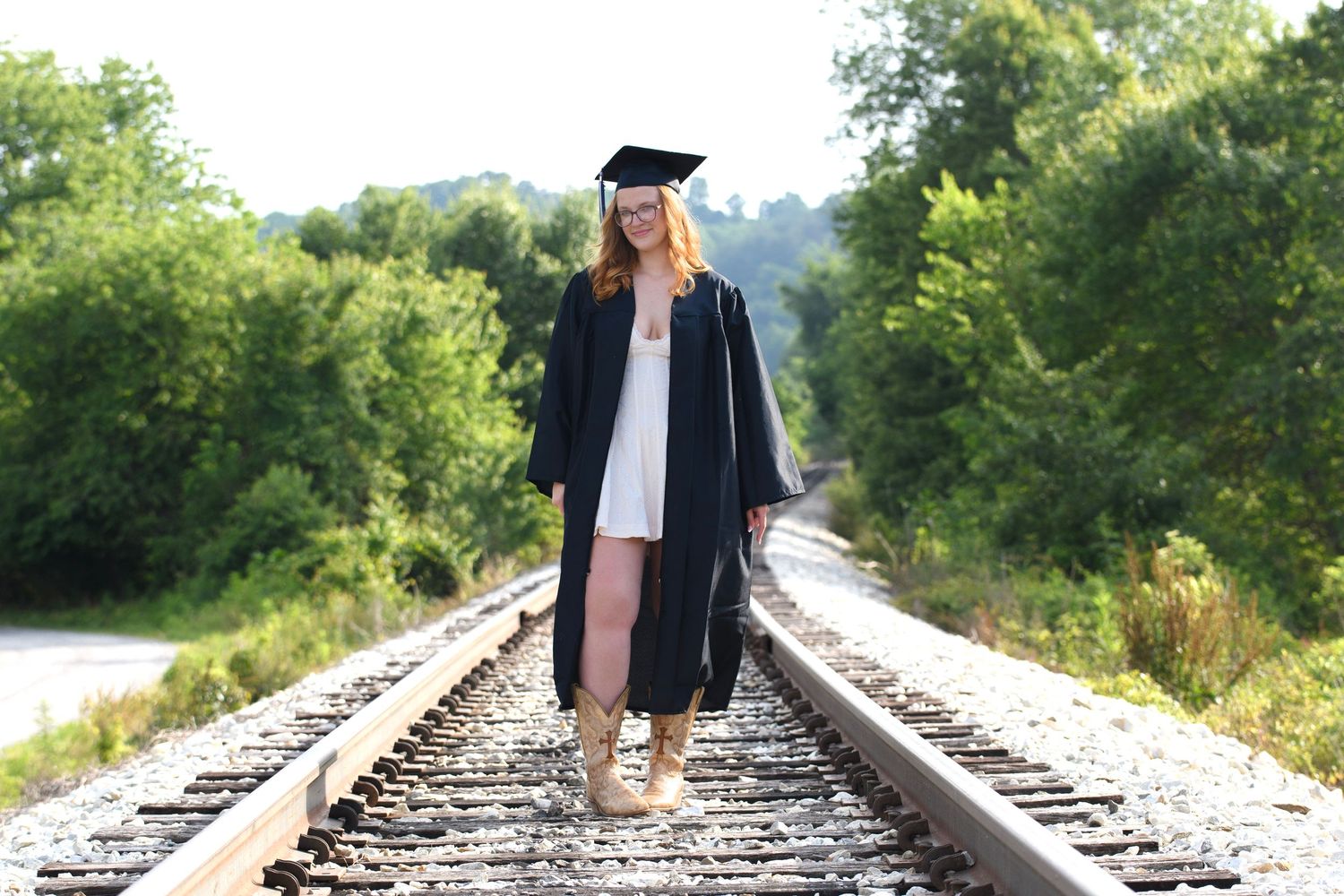Graduate in cap and gown walking on railroad tracks surrounded by lush green trees on a sunny day.