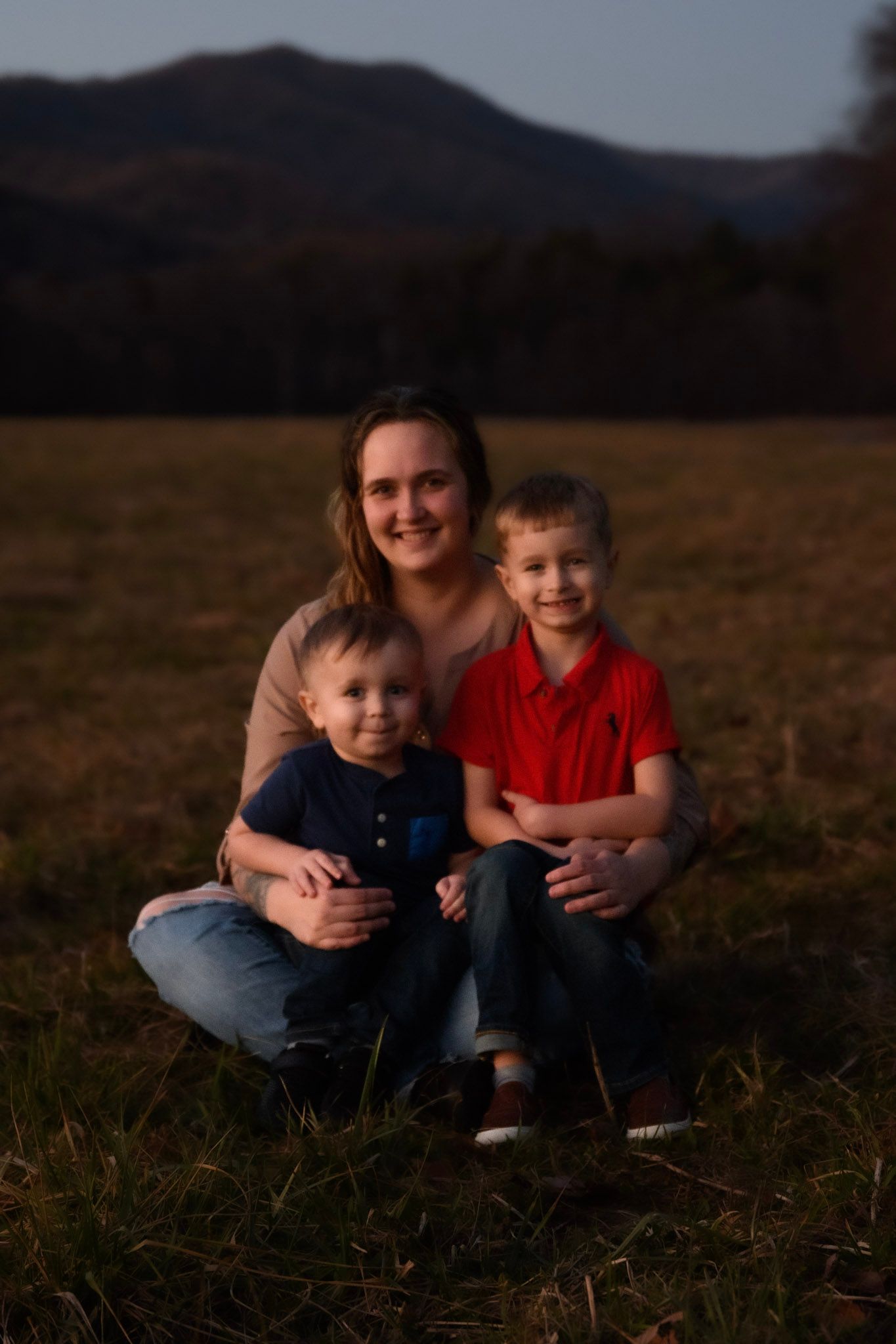 Smiling mother sitting with two young sons in a field at golden hour.