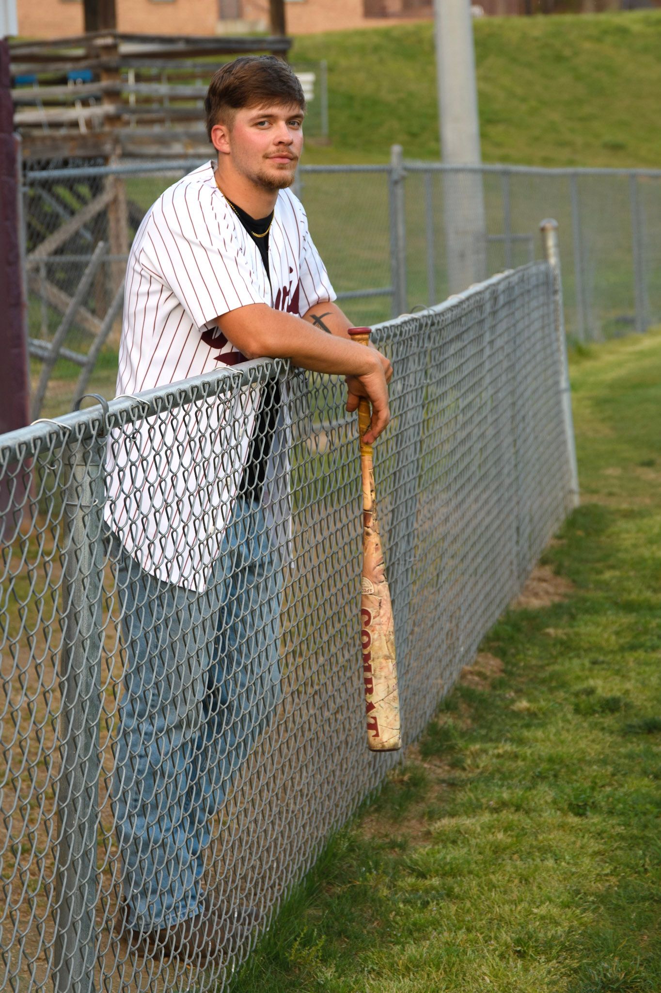 Young baseball player in pinstripe jersey leaning on chain-link fence holding a bat on a sunny day.