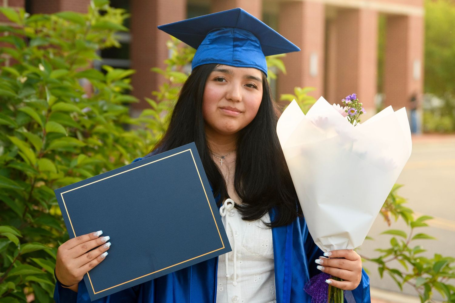 Young woman in blue graduation cap and gown holding diploma and flower bouquet outdoors.