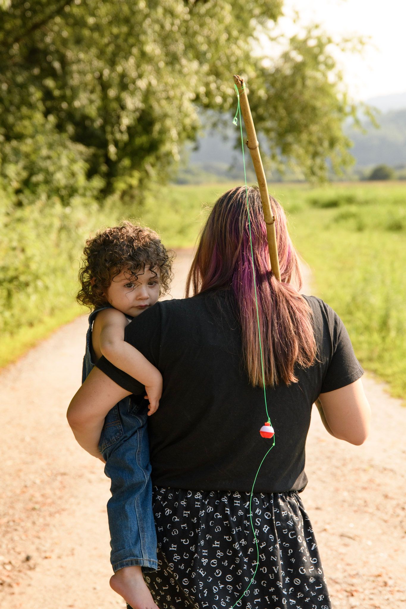A woman with colorful hair carries a curly-haired child while holding a fishing pole on a rural dirt path.