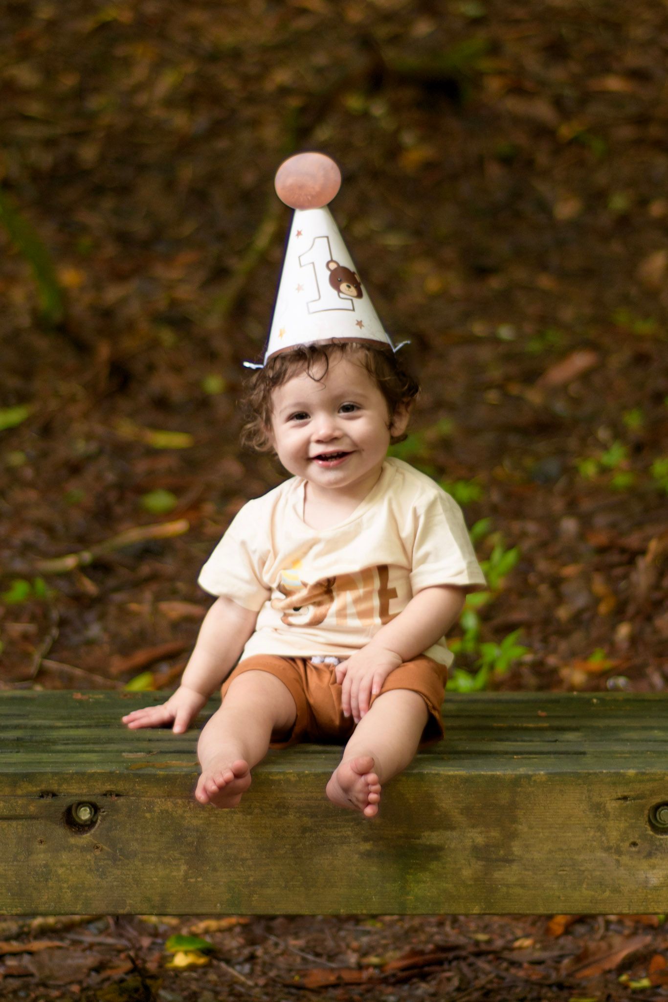 Happy toddler wearing a birthday party hat sitting on a wooden bench outdoors in a forest, smiling joyfully.