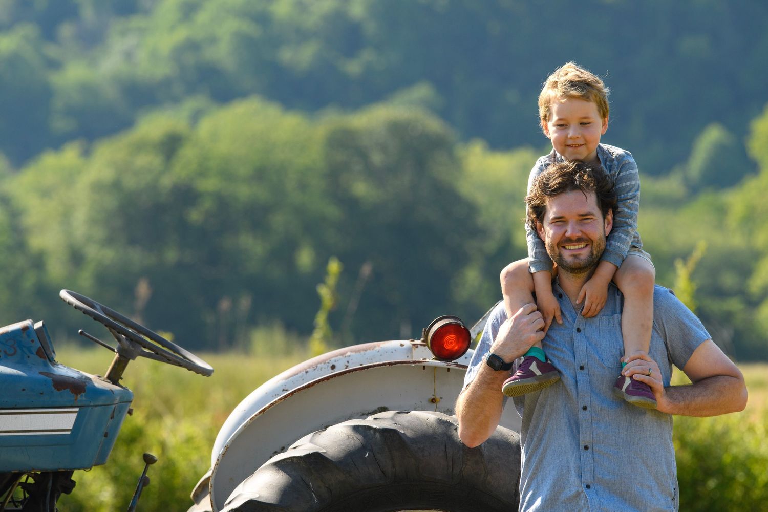 Smiling father with son on his shoulders standing beside a tractor in a green field.