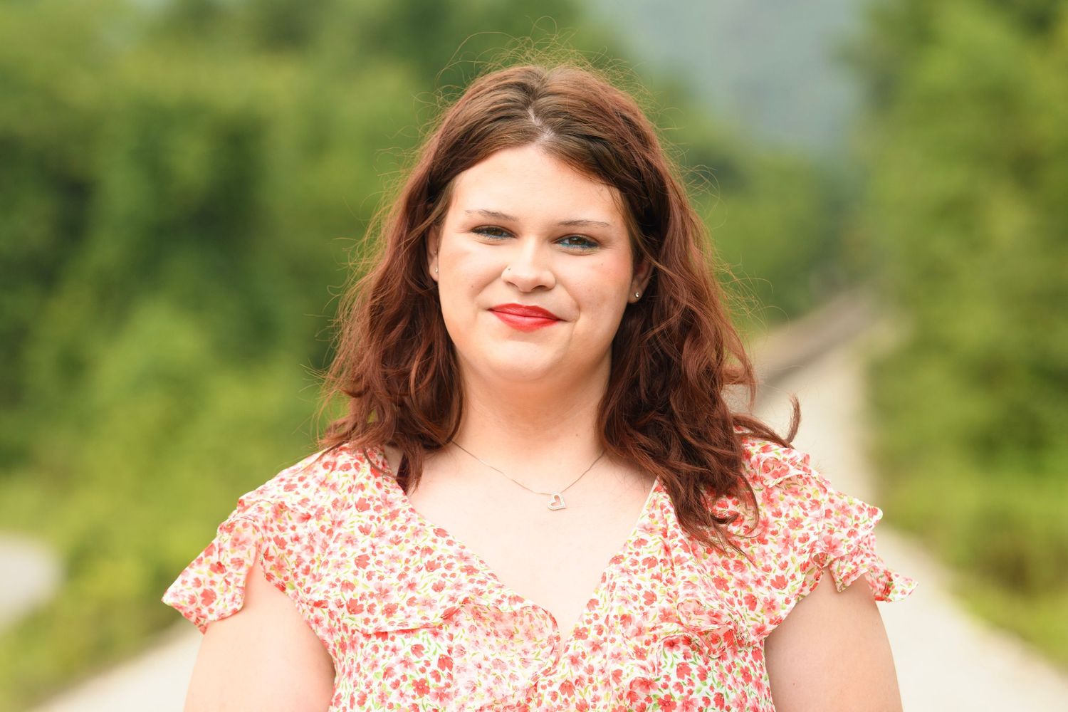 Young woman with auburn hair smiling outdoors in a floral pink dress with green trees in background.