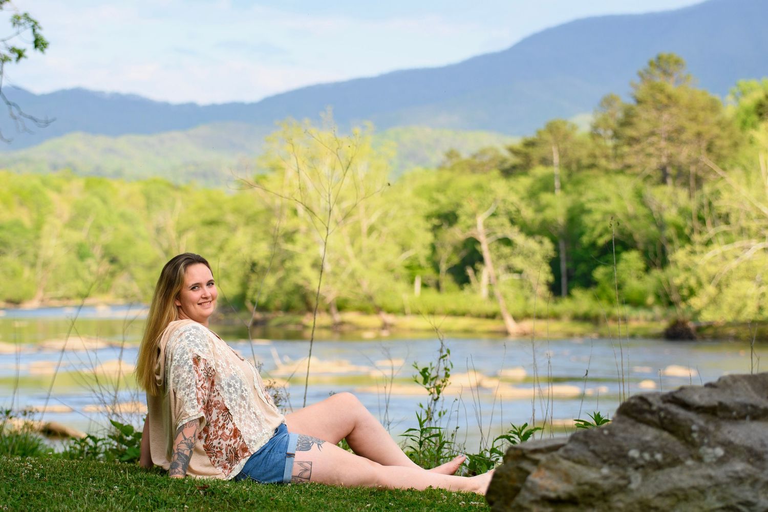 Smiling young woman in lace top sitting on grass beside a river with scenic mountain backdrop.