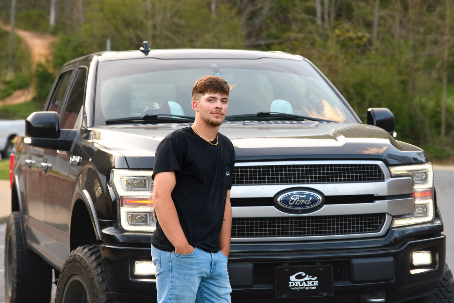 Young man in black t-shirt and jeans posing in front of a lifted Ford F-150 truck outdoors.