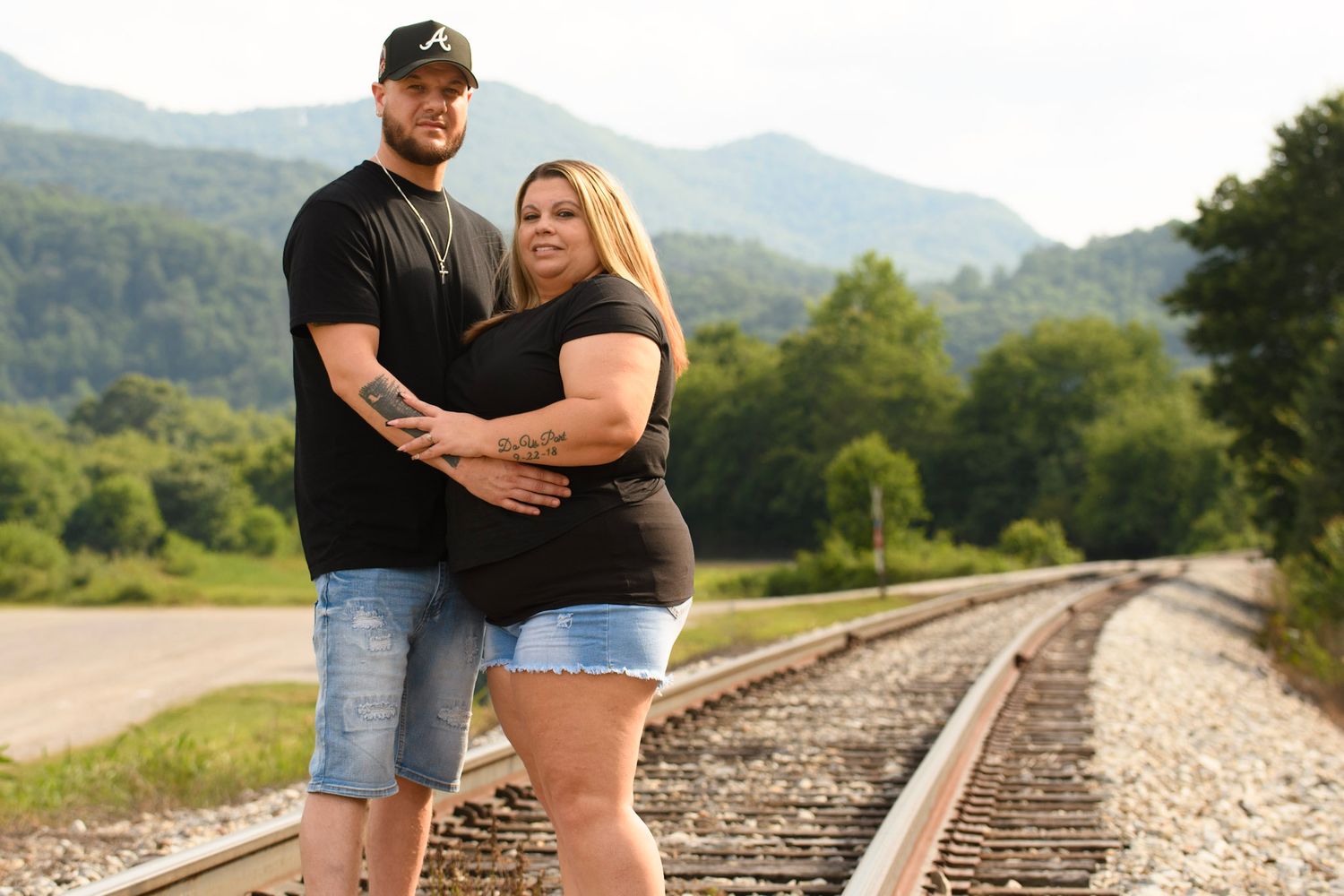 Couple posing on railroad tracks with green mountains in background on a sunny day.