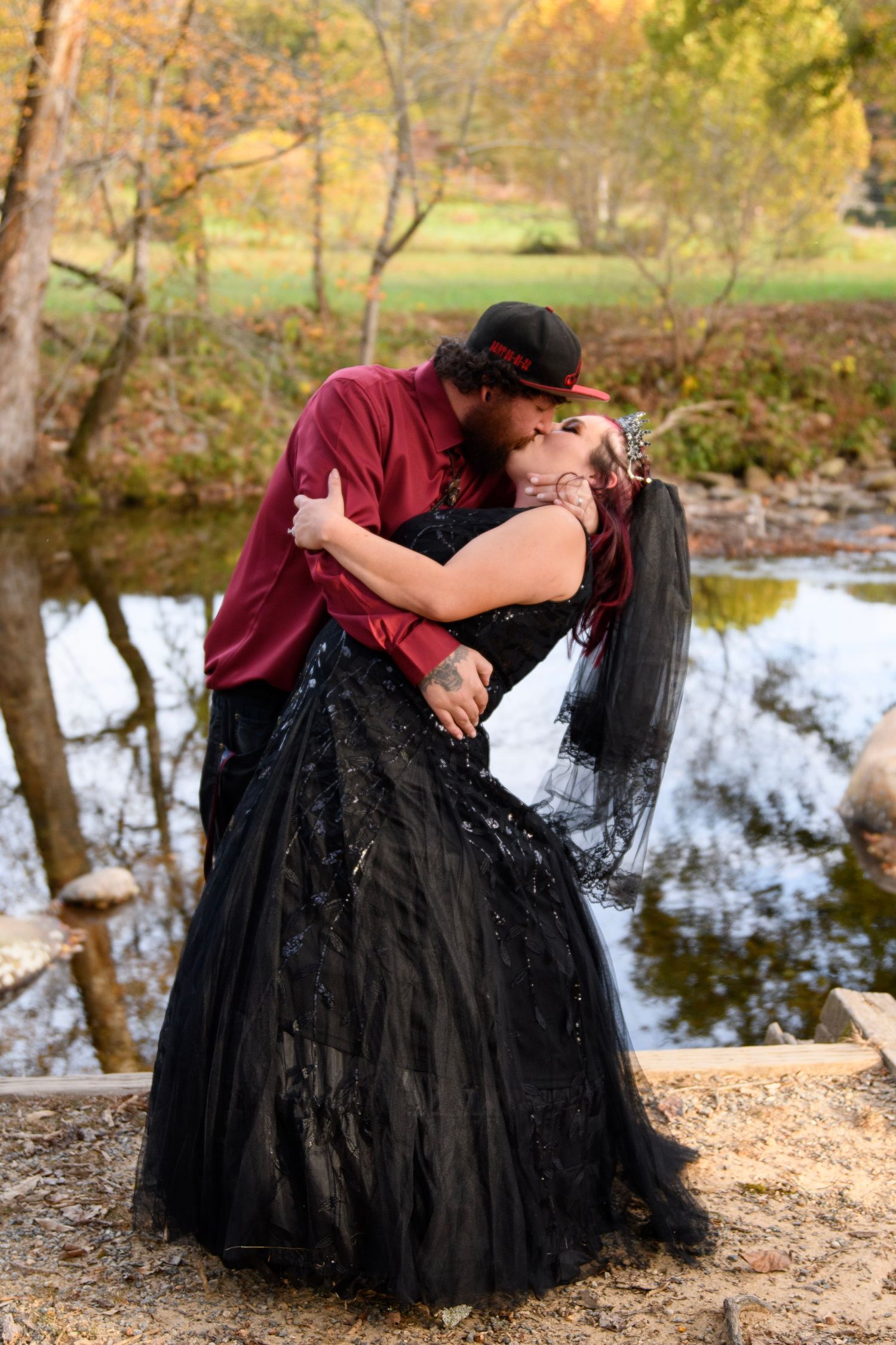 Couple sharing romantic kiss outdoors in autumn, bride in black gothic gown and groom in red shirt by a creek.