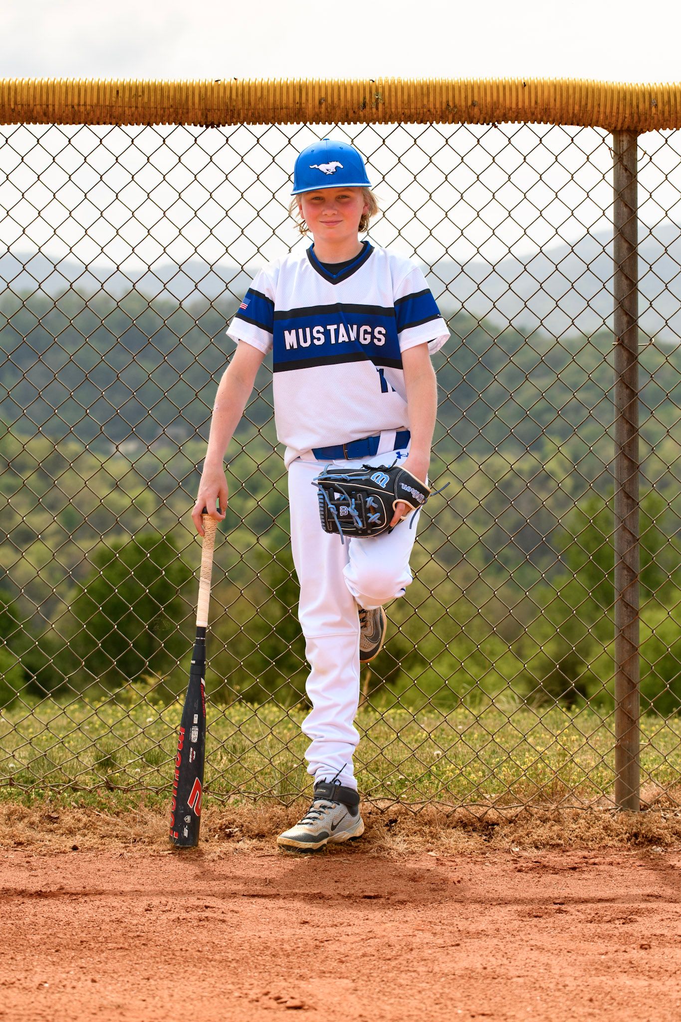 Young baseball player in Mustangs uniform holding bat beside chain-link fence on dirt field.
