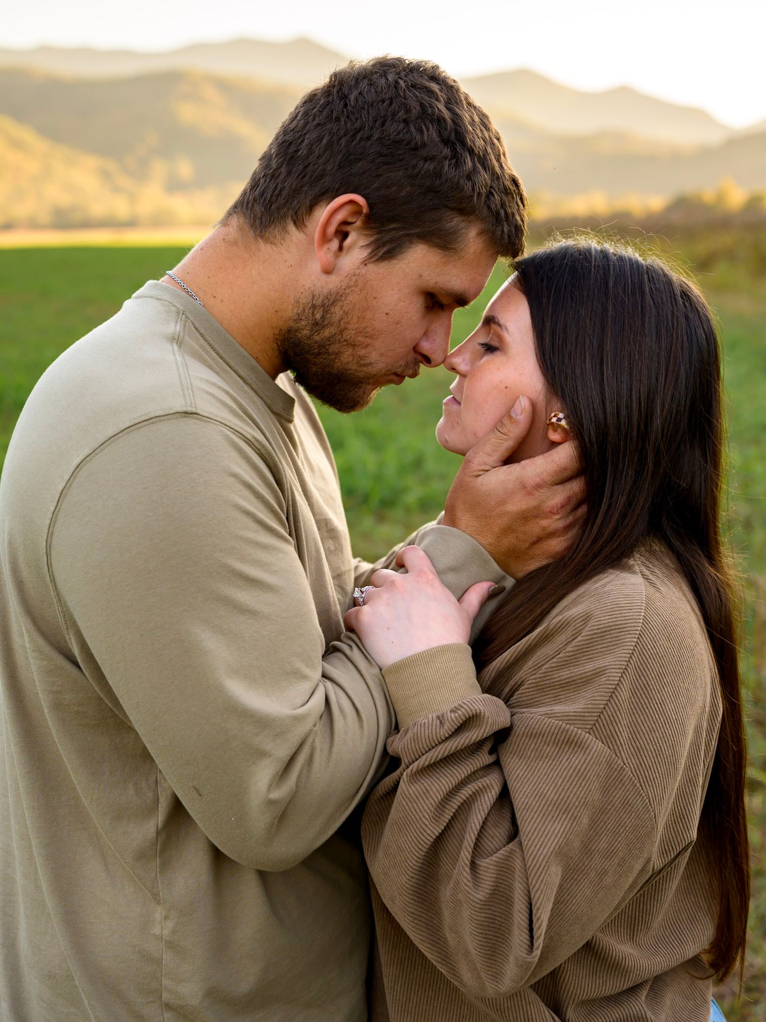 Romantic couple in earth-tone sweaters embrace with foreheads touching in a green field with golden mountain views at sunset.