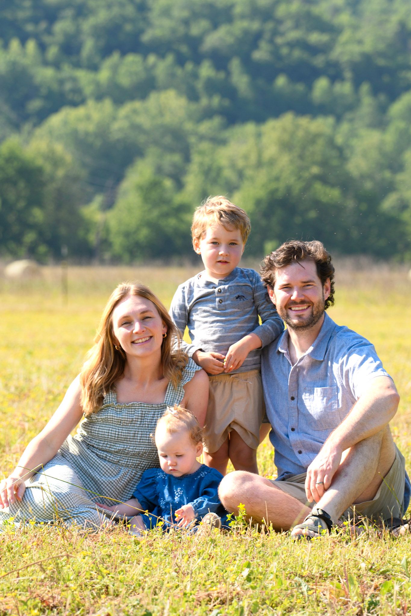 A happy family of four sitting in a sunny green field with wooded hills in the background.