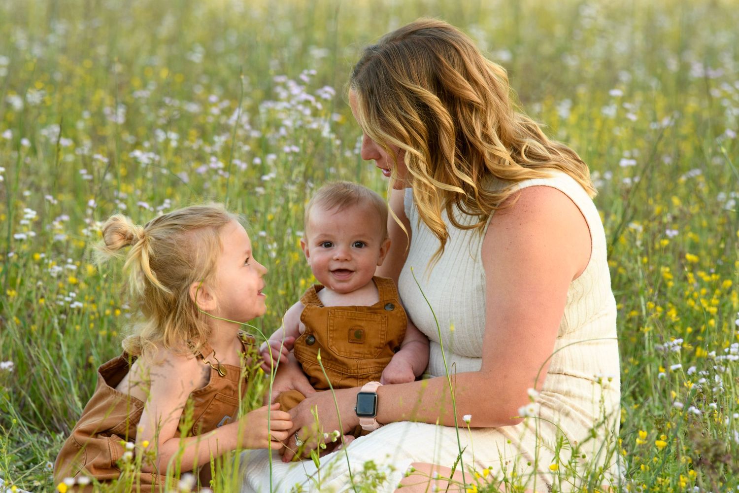 Mother sits in wildflower field holding baby while young daughter looks on adoringly.