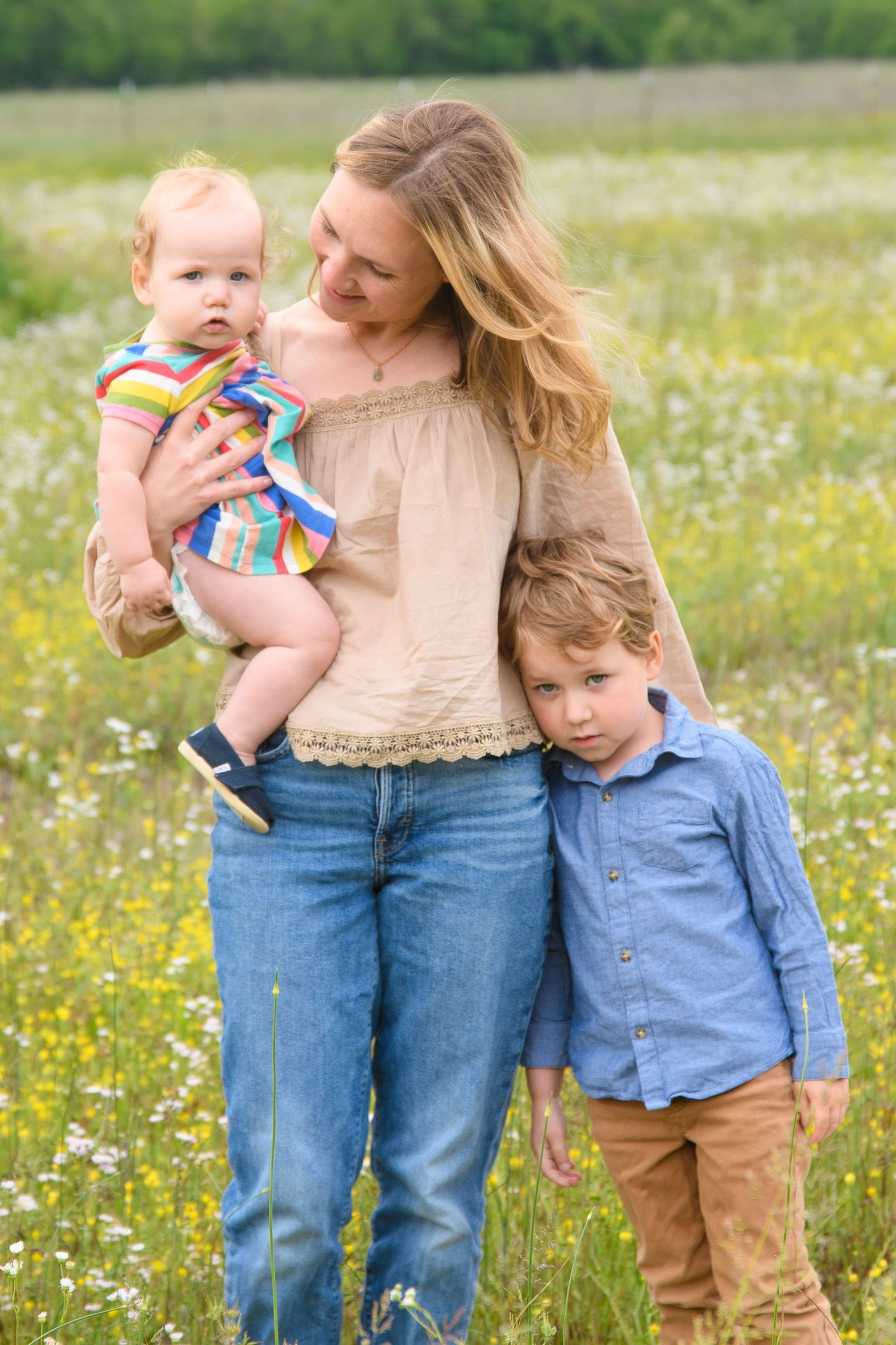 Mother holding baby in colorful outfit while young boy leans on her in a sunny wildflower field.