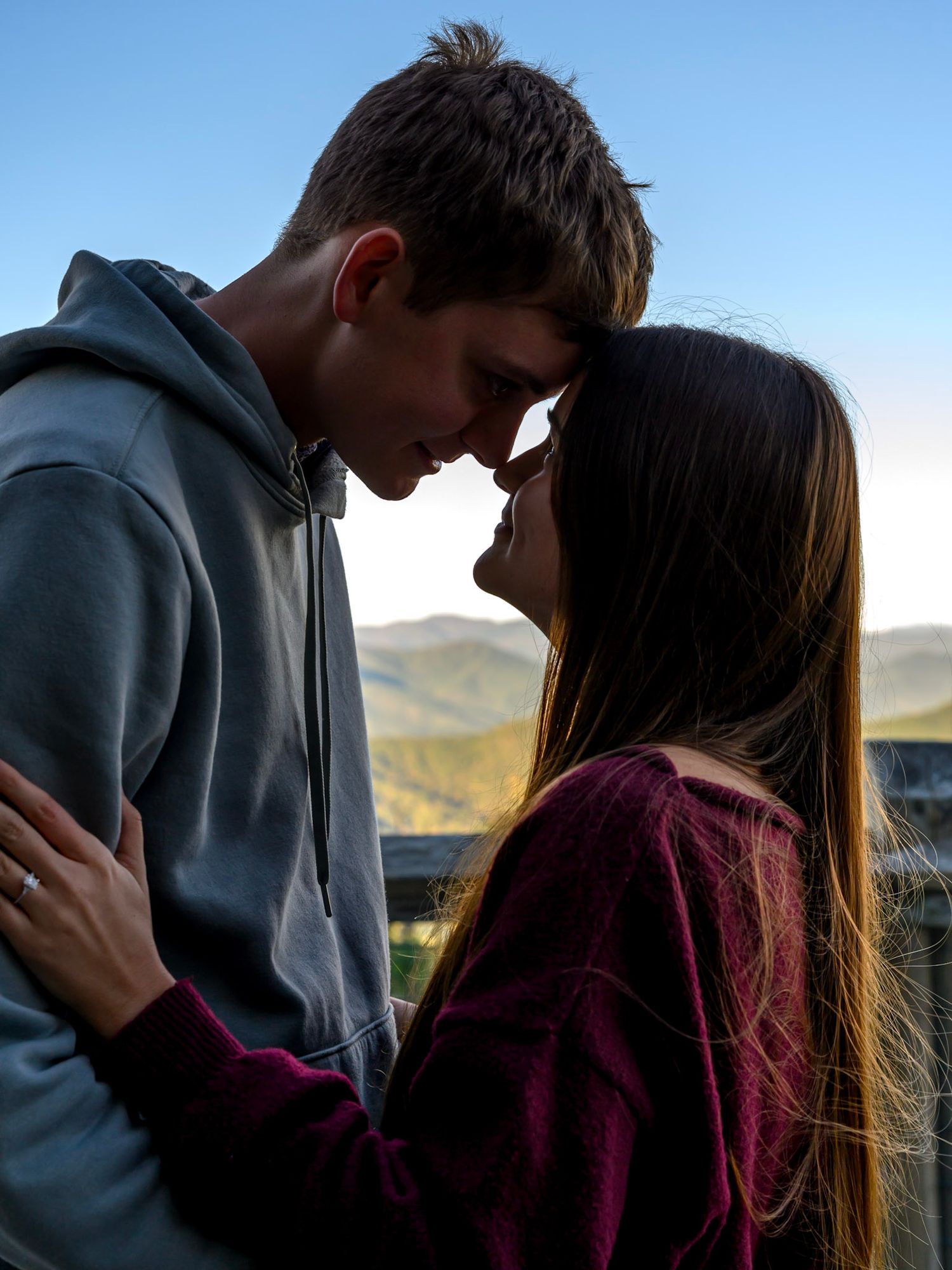 Young couple touching foreheads in a tender embrace on a mountain overlook with scenic hills and a clear blue sky behind them.