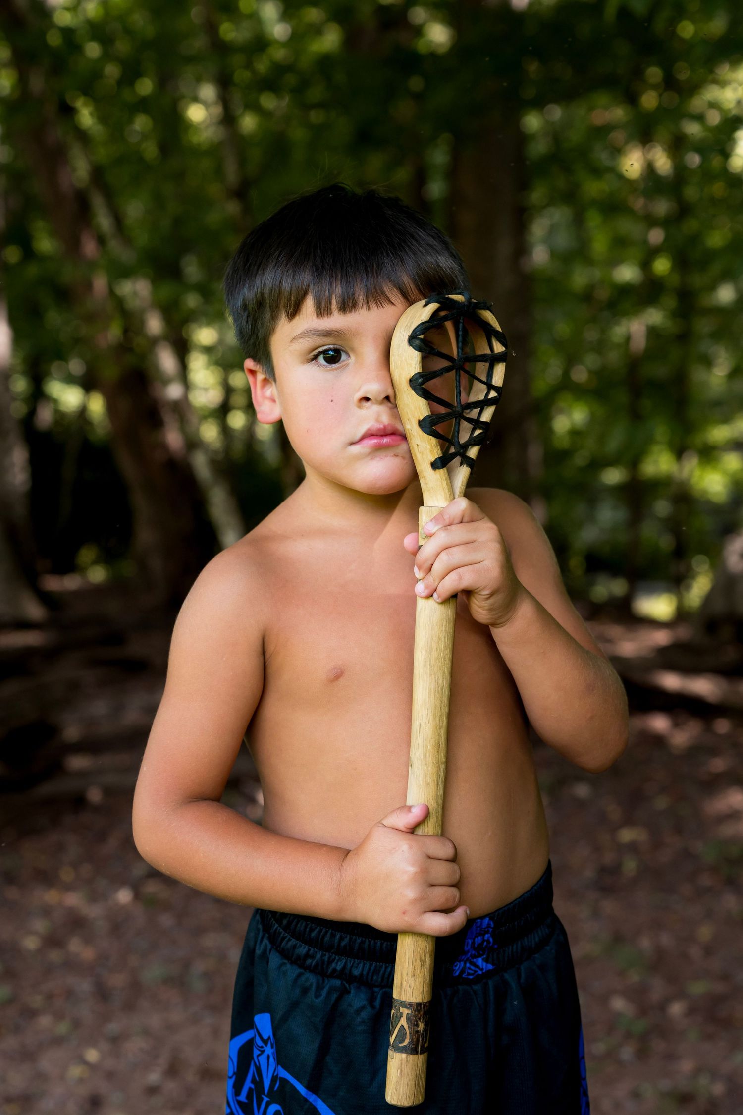 Shirtless young boy holding a wooden lacrosse stick in a forest setting, looking seriously at the camera.
