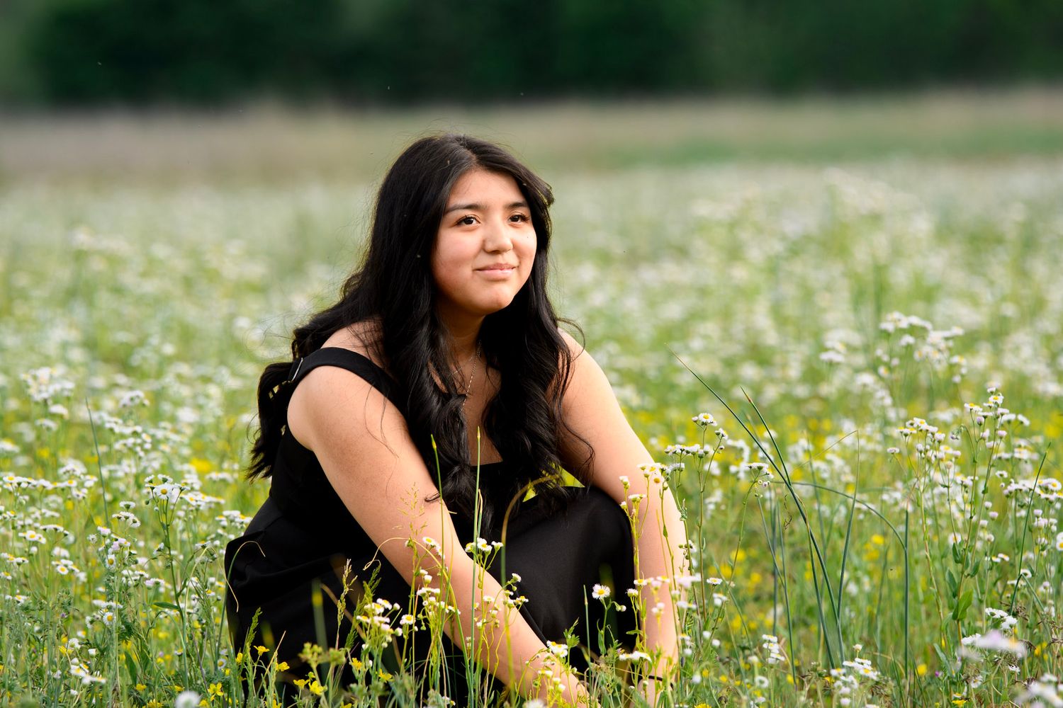 Young woman with long dark hair sits among wildflowers in a green meadow wearing a black outfit.