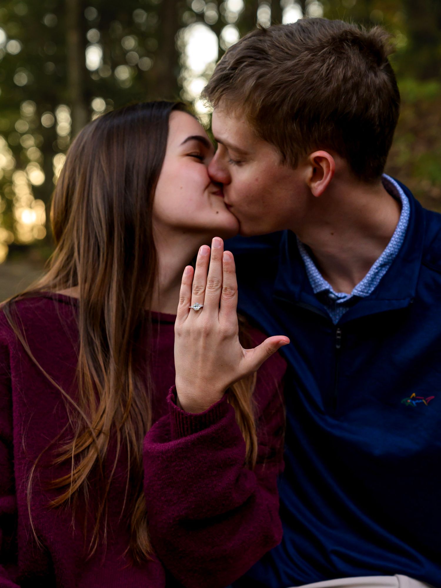 A couple shares a kiss in a forest while the woman holds up her hand to show her new engagement ring.