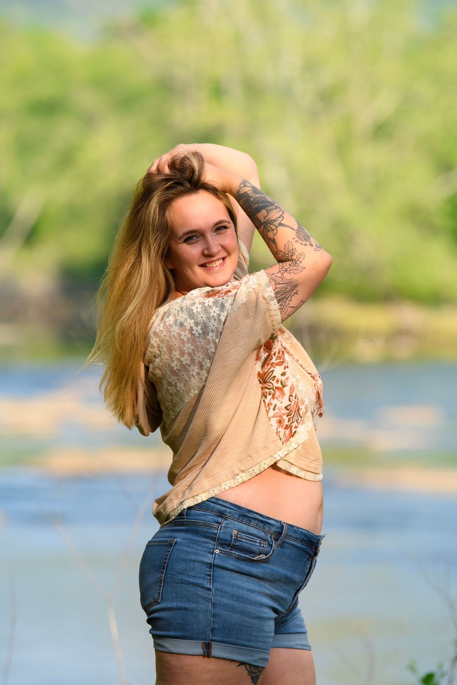 Smiling blonde woman with tattoo posing in denim shorts near scenic river.