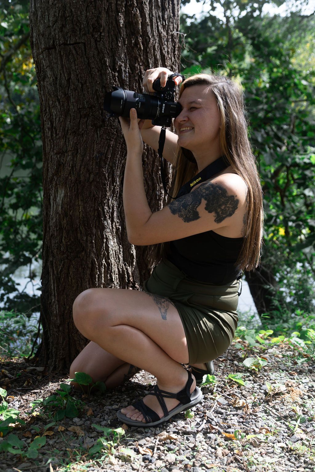 Woman with tattoos crouching by a tree in nature, smiling while holding a DSLR camera outdoors.