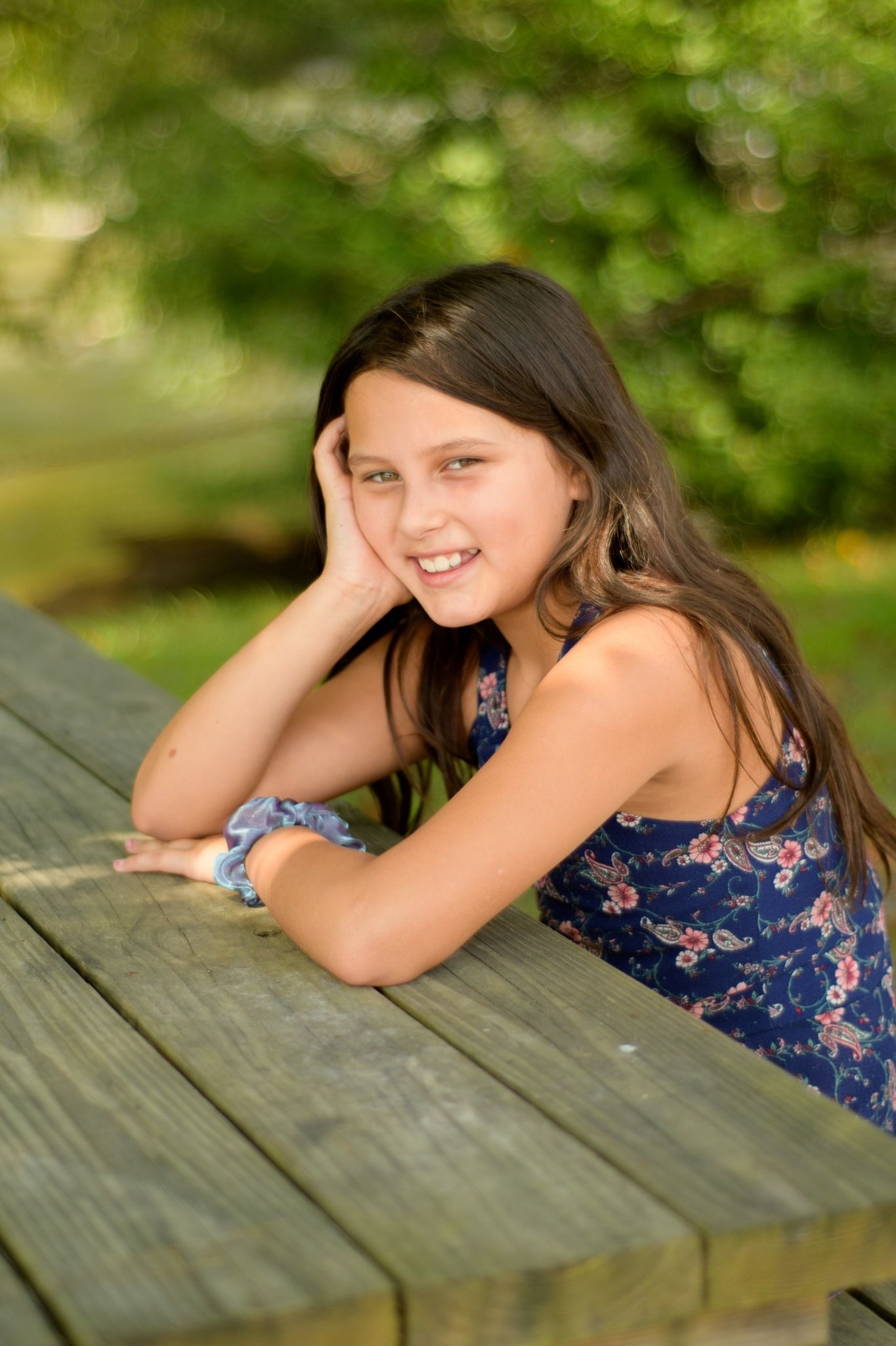 Smiling young girl with long brown hair leaning on a wooden picnic table outdoors.
