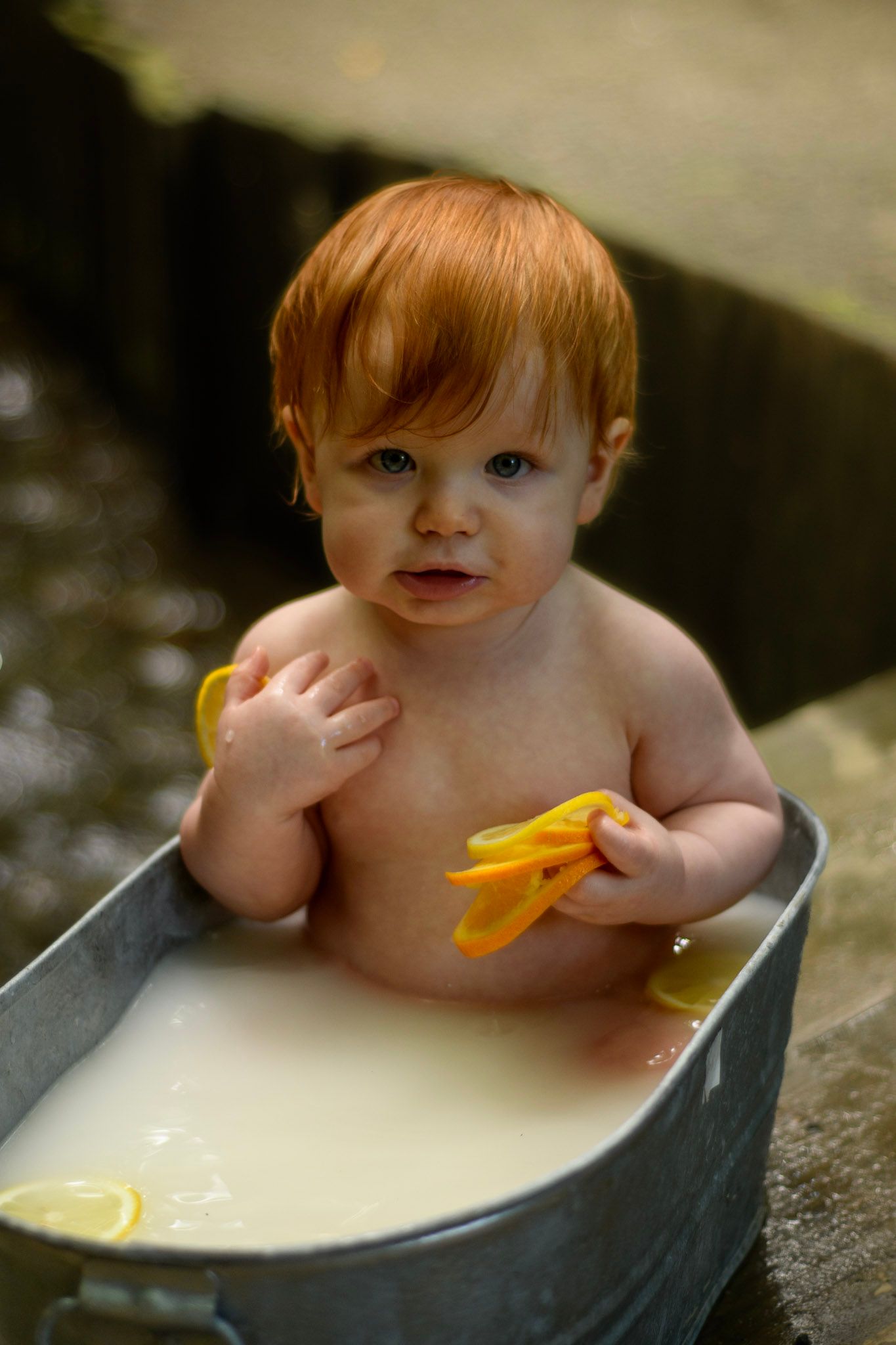 Adorable red-haired baby sitting in metal tub with milk bath holding yellow rubber toy.